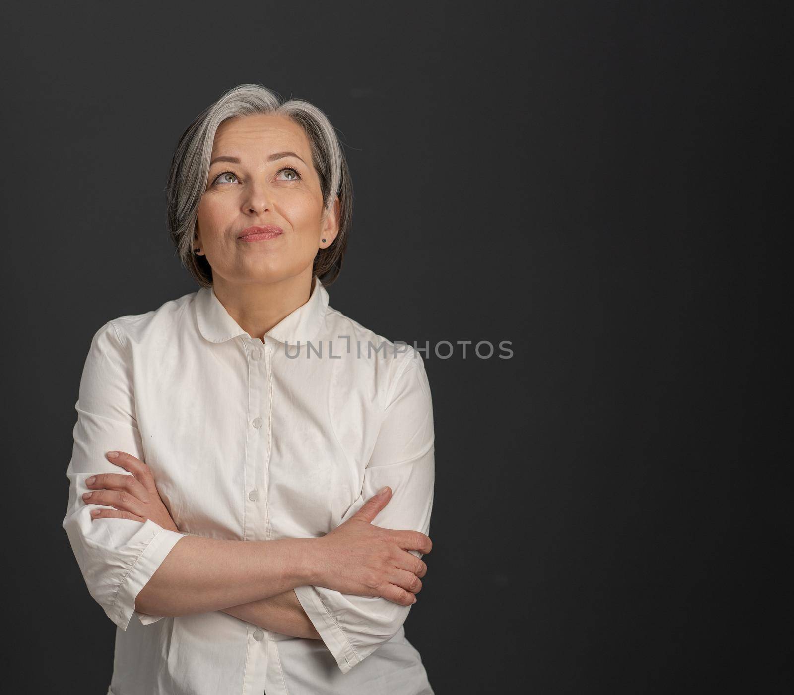 Creative mature woman looking upward thoughtfully with her arms crossed. Charming silver-haired woman smiling tenderly on dark grey background with textspace at right side.