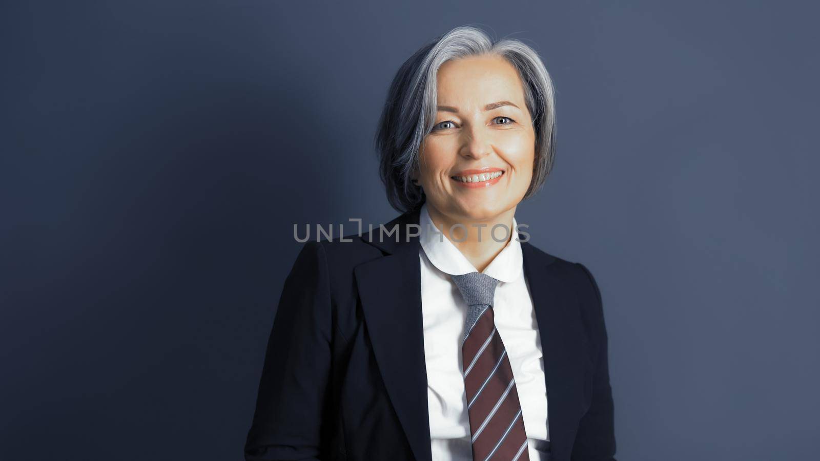 Positive middle-aged businesswoman without mask smiles happily. Optimistic graying woman wearing formalwear posing on blue gray background. Text or Copy space on both sides. Studio shot. Toned image.