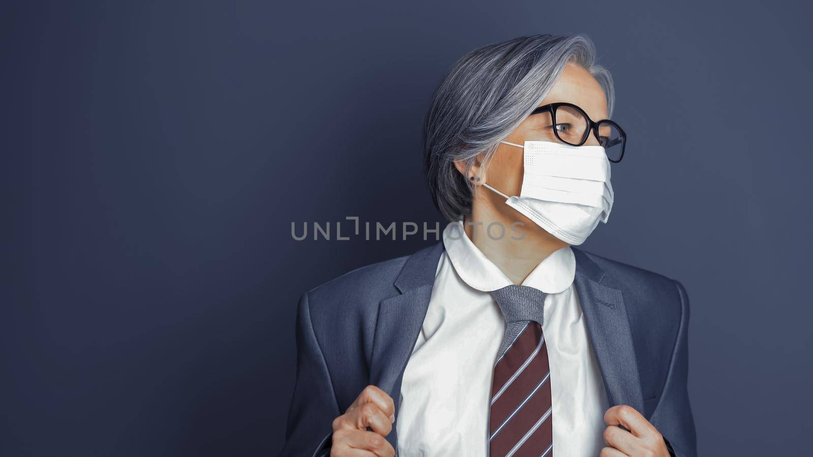 Mature woman wearing mask corrects her jacket looking at right side. Caucasian smart business woman on gray background. Copy space at left. Quarantine concept. Toned image by LipikStockMedia