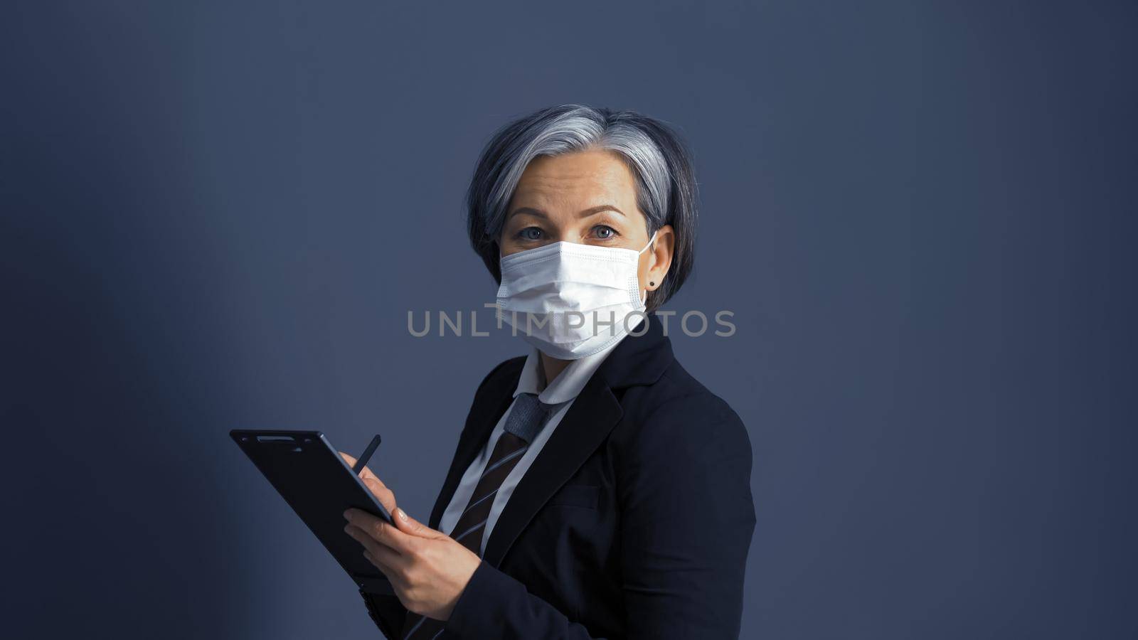 Confident middle-aged businesswoman in protective mask using digital notepad. Graying female model looks at camera, isolated on gray background in studio. Toned image by LipikStockMedia