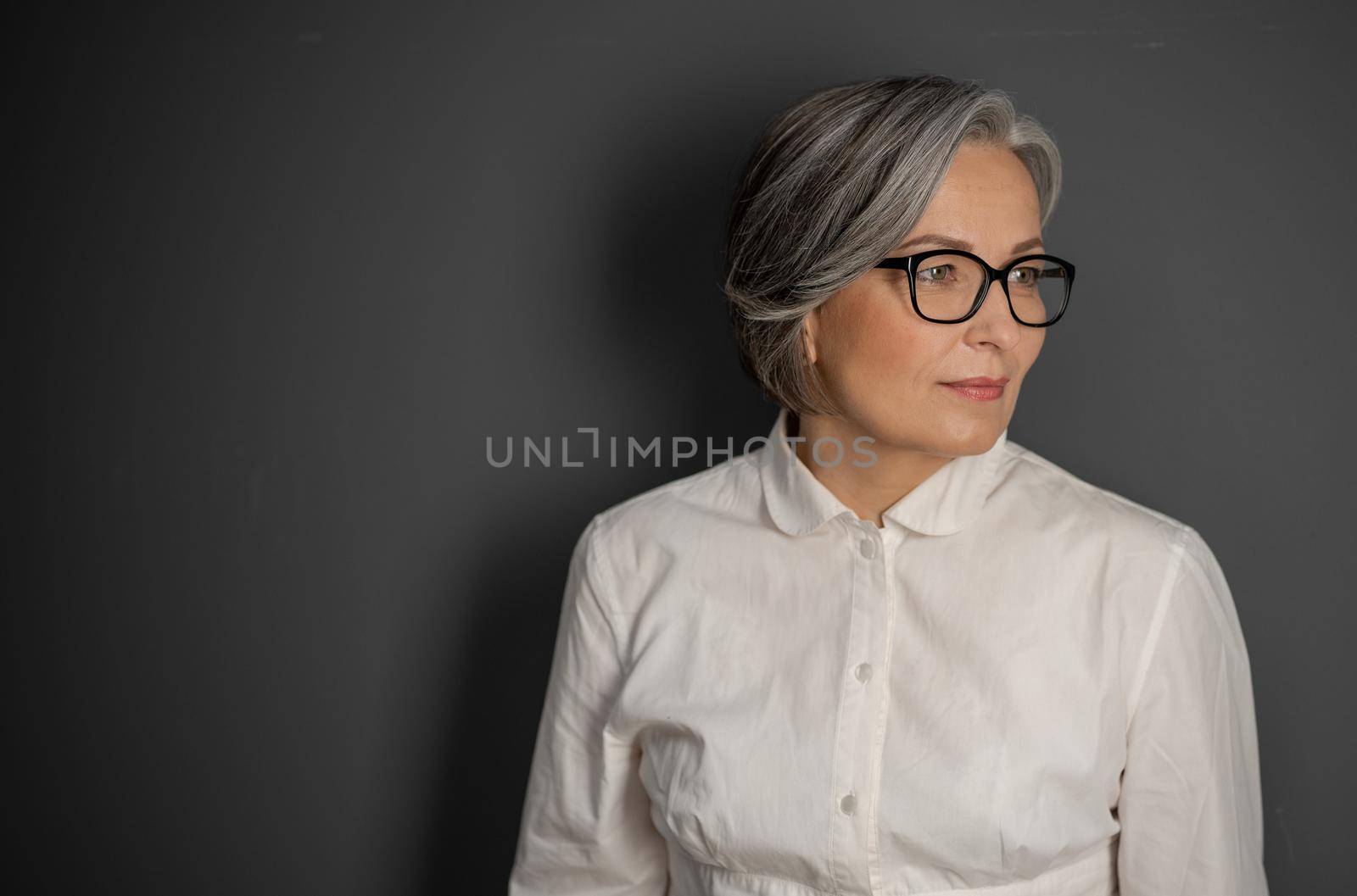 Beautiful gray-haired business woman looking at side. Mature woman in glasses and white shirt on grey background. Copyspace at left side.