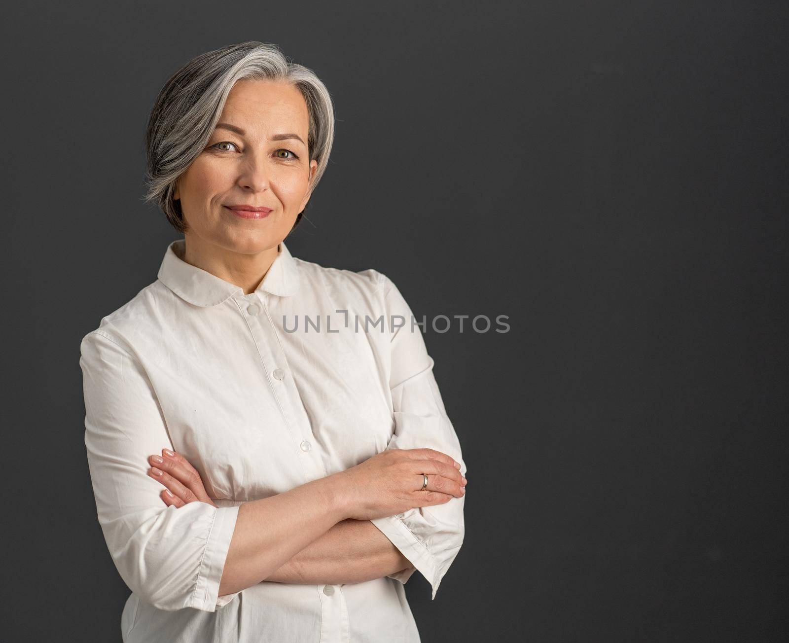 Attractive gray-haired middle-aged woman smiles looking at camera with crossed arms. Copy space at right side on gray background.