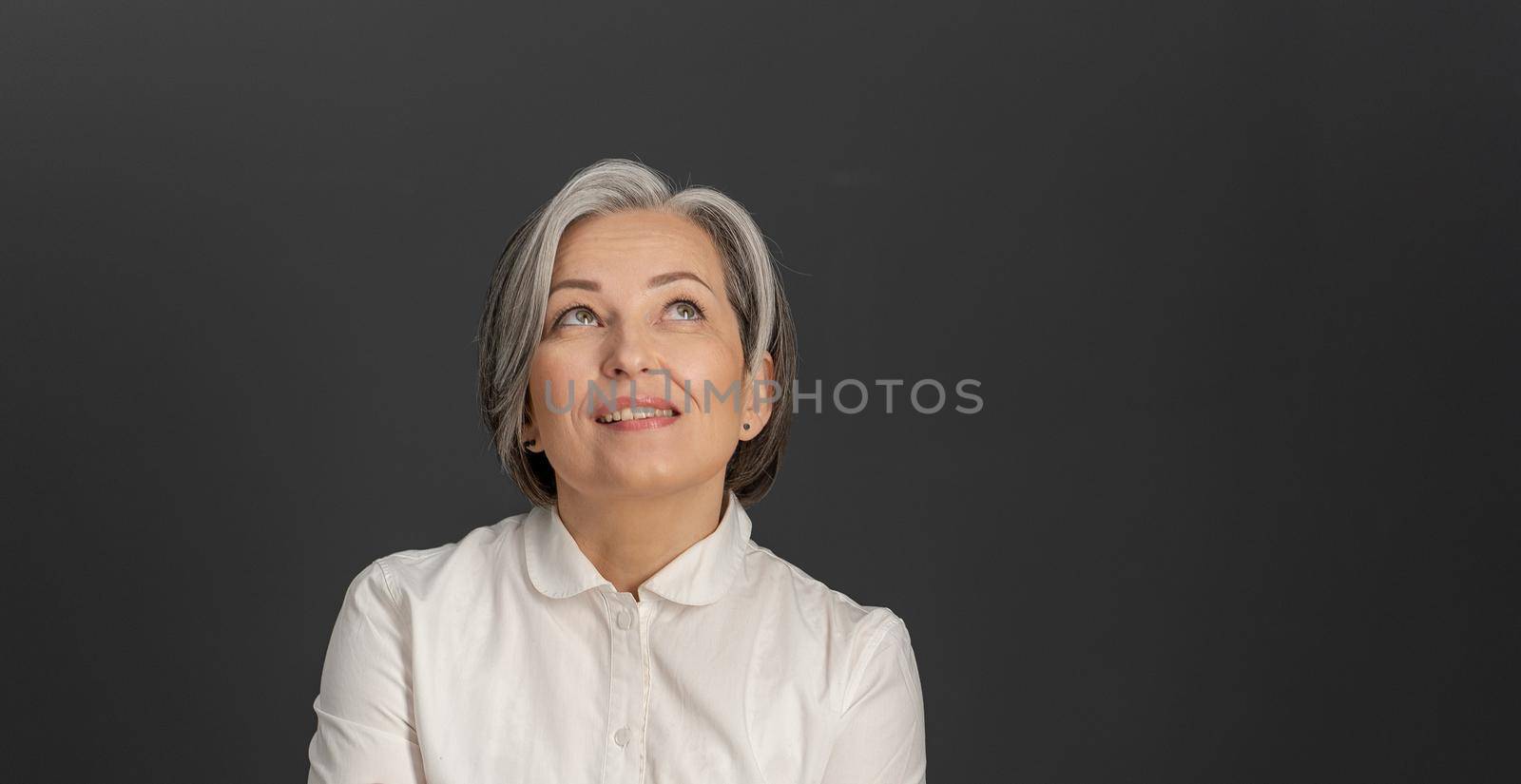 Thinking white-haired business woman looking upward. Creative middle-aged pretty woman thoughtfully smiles posing on grey background. Close up portrait. Copyspace at right side.