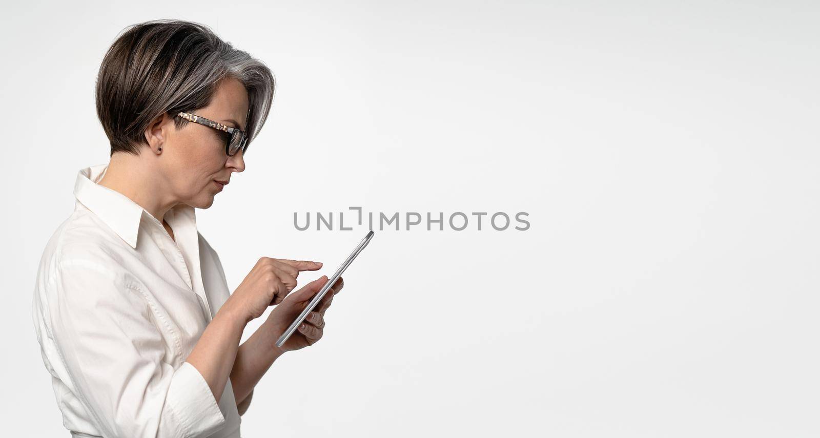 Gray-haired Caucasian businesswoman typing on tablet computer on white background. Horizontal template for ad banner with textspase at right by LipikStockMedia