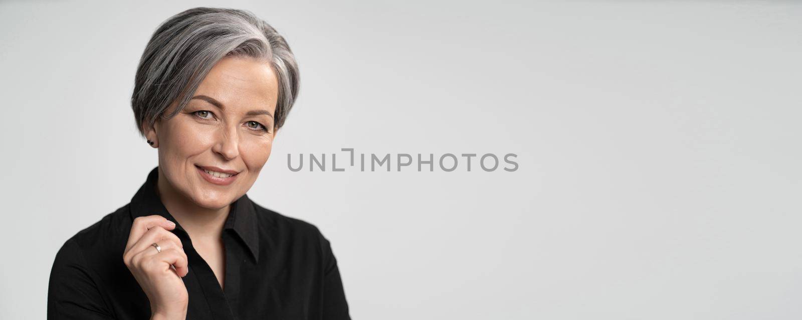 Pretty mature woman smiles with hand near face. Charming gray-haired lady in black blouse on white background. Copy spase for text. Horizontal blank or template for ad banner.