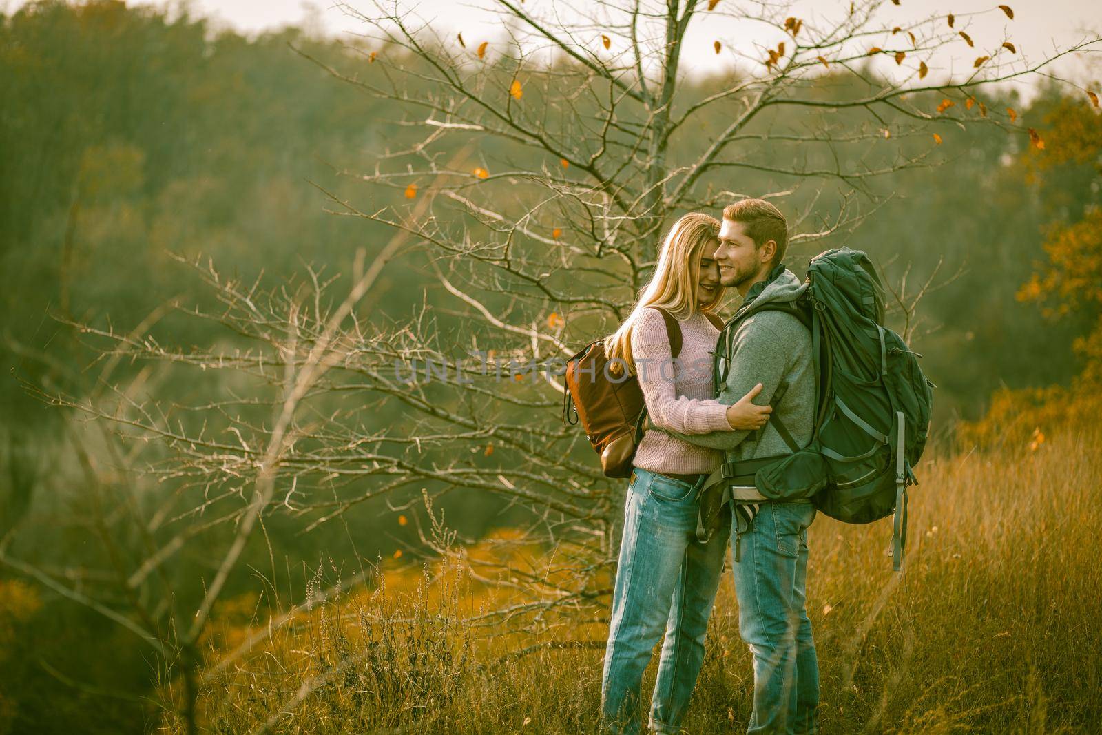 Happy couple admires of nature standing with backpacks on autumn grass. Young Caucasian man and woman smile embracing while enjoying the beautiful view. Togetherness with nature concept.