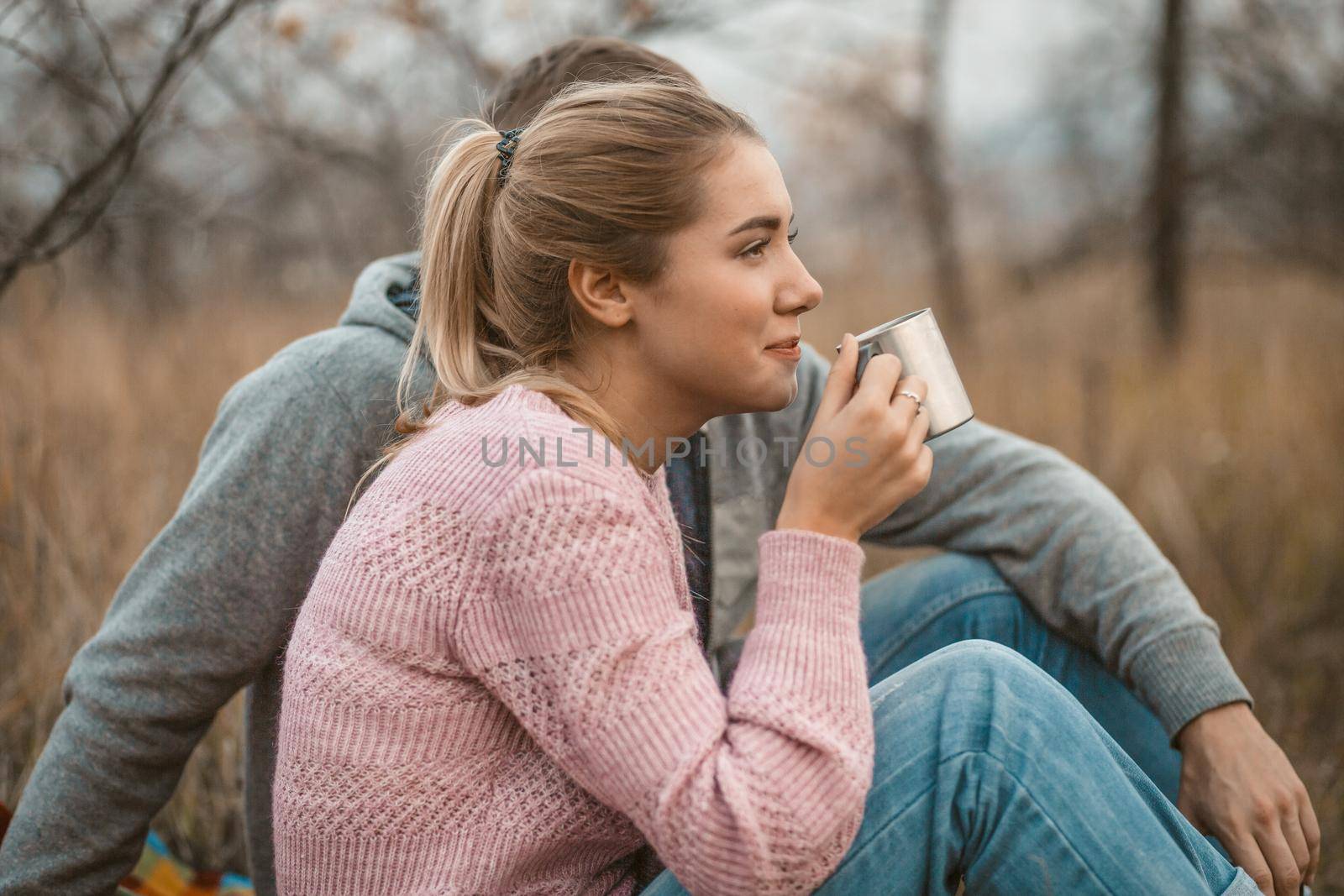 Picnic in nature. Young woman and man rests drinking hot coffee or tea while sitting on blanket at autumn grass outdoors. Profile view. Lifestyles concept.