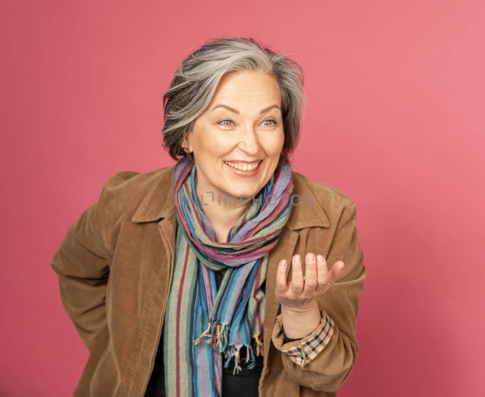 Funny mature woman laughs gesturing by hand and looking at side. Creative aged lady posing in studio on pink background.