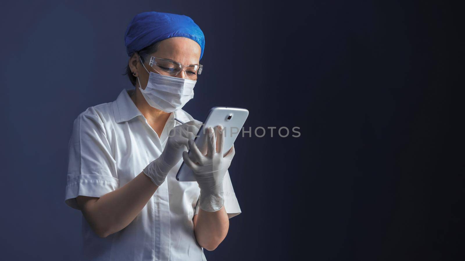 Doctor working with database on digital tablet. Caucasian woman in mask and gloves uses digital gadget standing on blue gray background with copy space for text on right side. Tinted image.