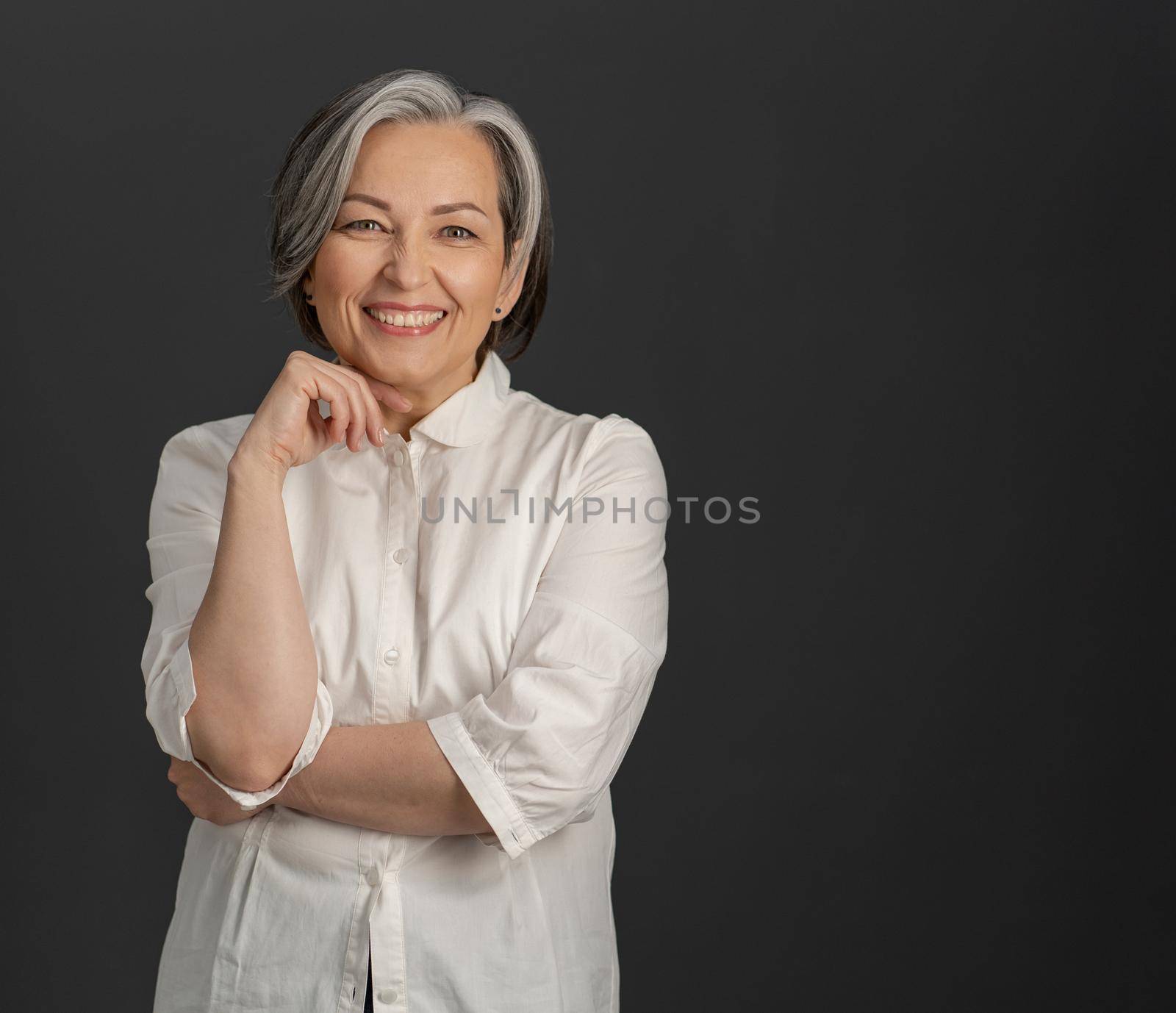 Happy gray-haired businesswoman toothy smiles at camera. Beautiful woman in whiite shirt posing on gray background with textspace at right by LipikStockMedia