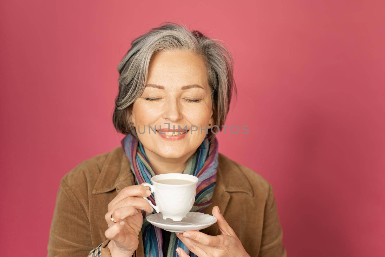 Charming mature woman enjoys by cup of coffee closing her eyes. Happy gray-haired lady drinks coffee posing on pink background in the studio. Close up portrait with text space on right.