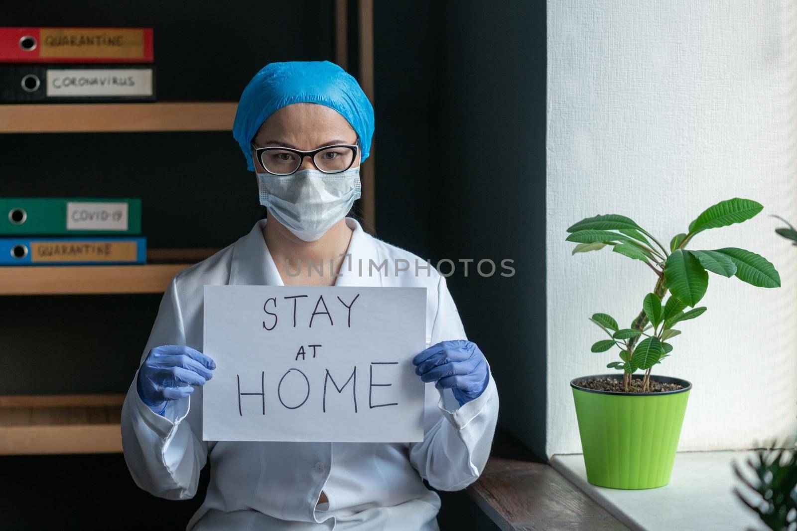 Doctor Shows Sign STAY AT HOME, Woman Wearing Protective Mask And White Medical Uniform Standing In The Office Next To The Window, Quarantine Concept