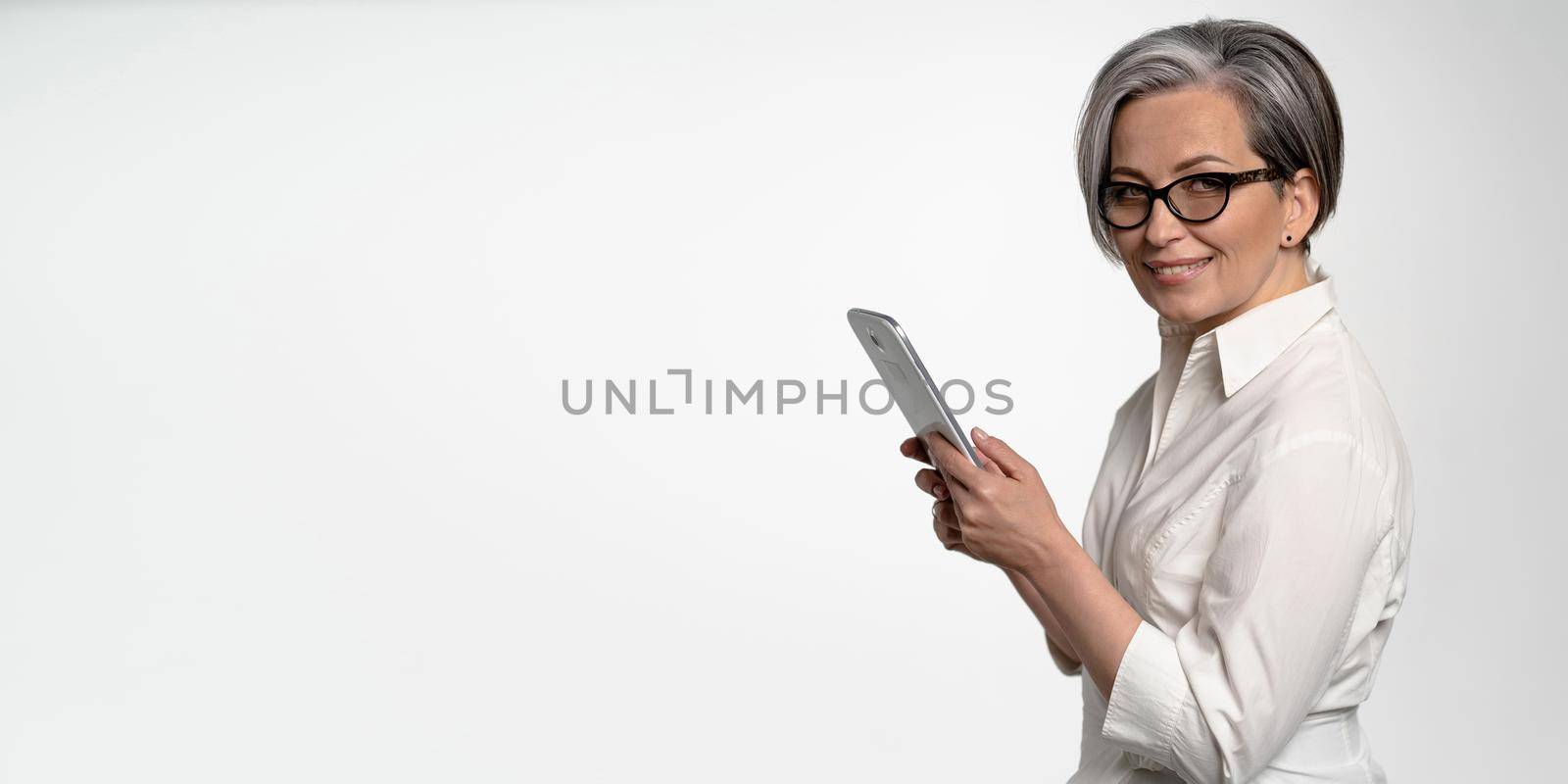 Cheerful business woman works on digital tablet on white background. Mature lady in white shirt looking at camera. Horizontal blank with empty spase for text at left side. Business or Education concept.