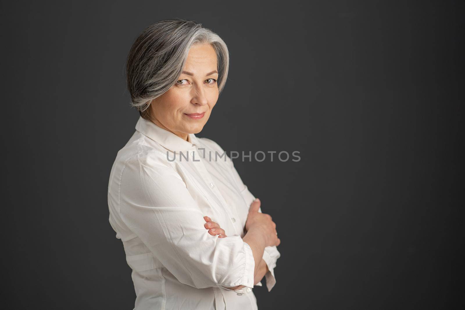 Pretty white-haired woman smiles looking at camera with arms crossed. Stylish caucasian businesswoman isolated on dark gray background. Textspace at right side. Business concept.