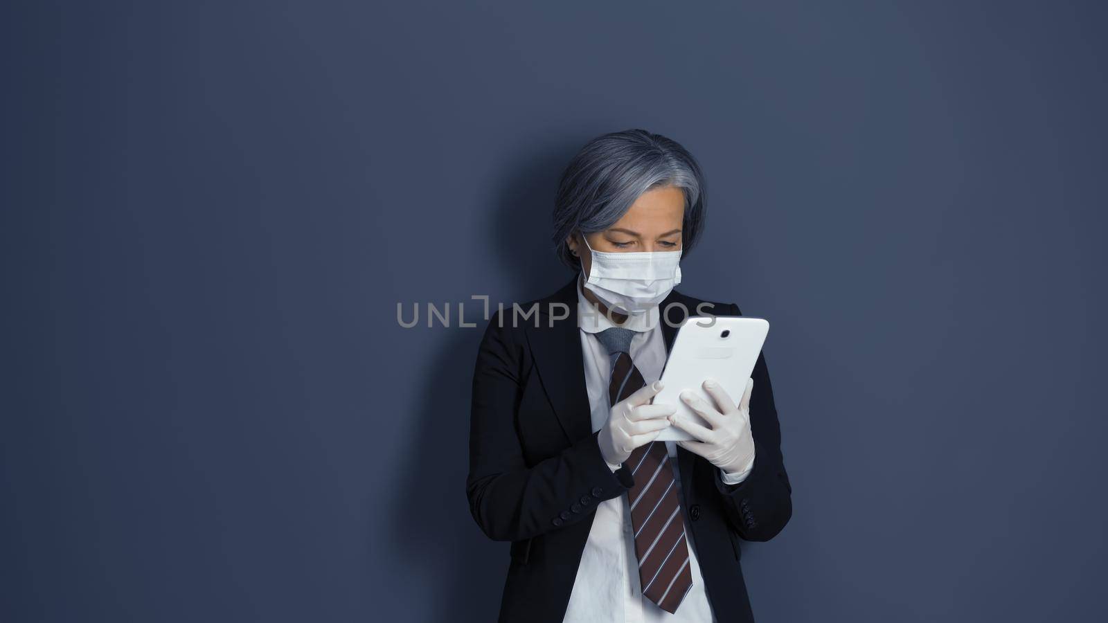 Mature woman works with digital tablet. Gray-haired businesswoman in black suit using electronic gadget standing against on gray office wall.