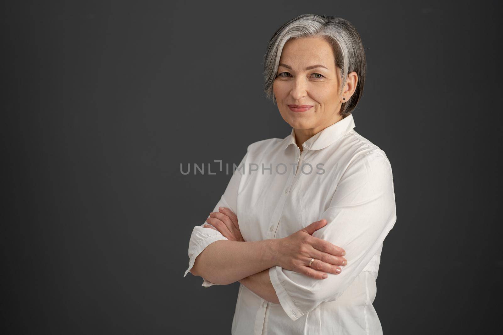 Intelligent silver-haired woman smiles with arms crossed. Pretty business woman looking at camera confidently. Isolated on gray background. Copyspace at left side. Success concept.