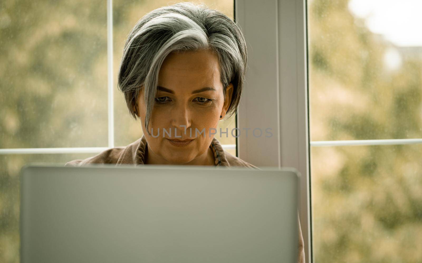 Mature woman works laptop sitting on sill. Serious Caucasian Lady looking at computer monitor against window. Front view. Close up shot. Tinted image by LipikStockMedia