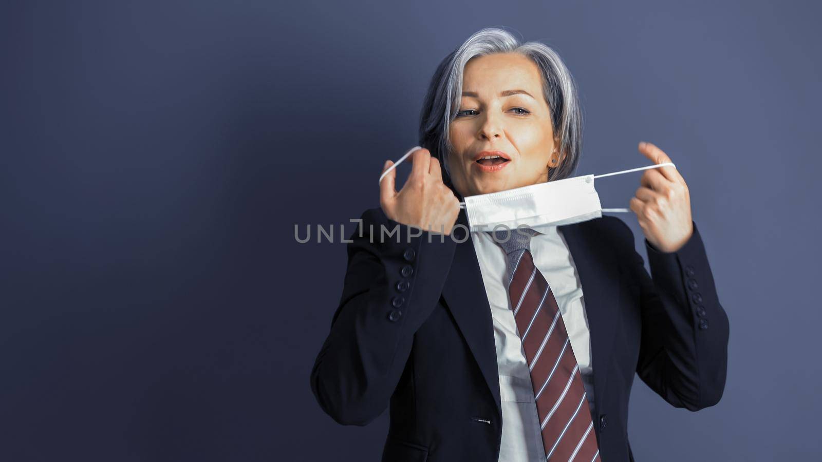 Business woman removes protective mask from her face. Focus on female face. Mature woman in formalwear on blue gray background. Copy space at left side. Close up portrait. Tinted image.