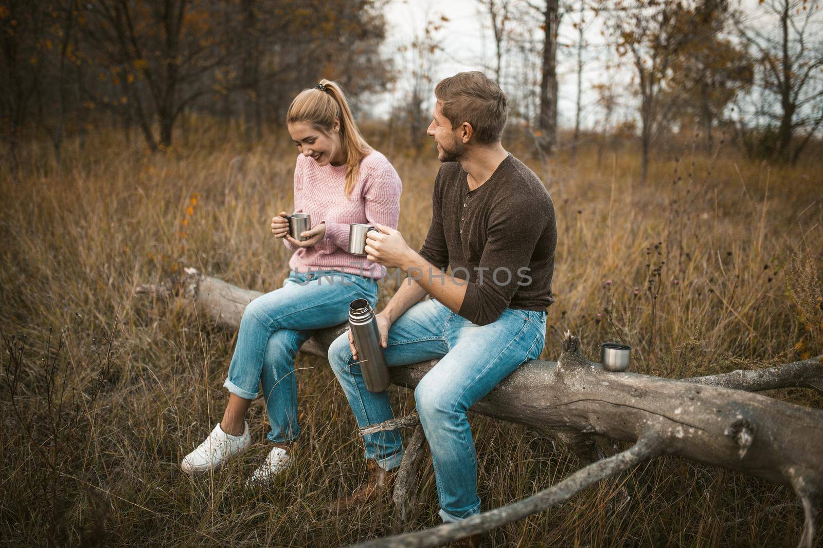 Couple of tourists drink hot drink in nature. Smiling Caucasian man and woman have rest outdoors holding metal camping mugs with hot coffee or tea from thermos sitting on old tree among forest lawn.