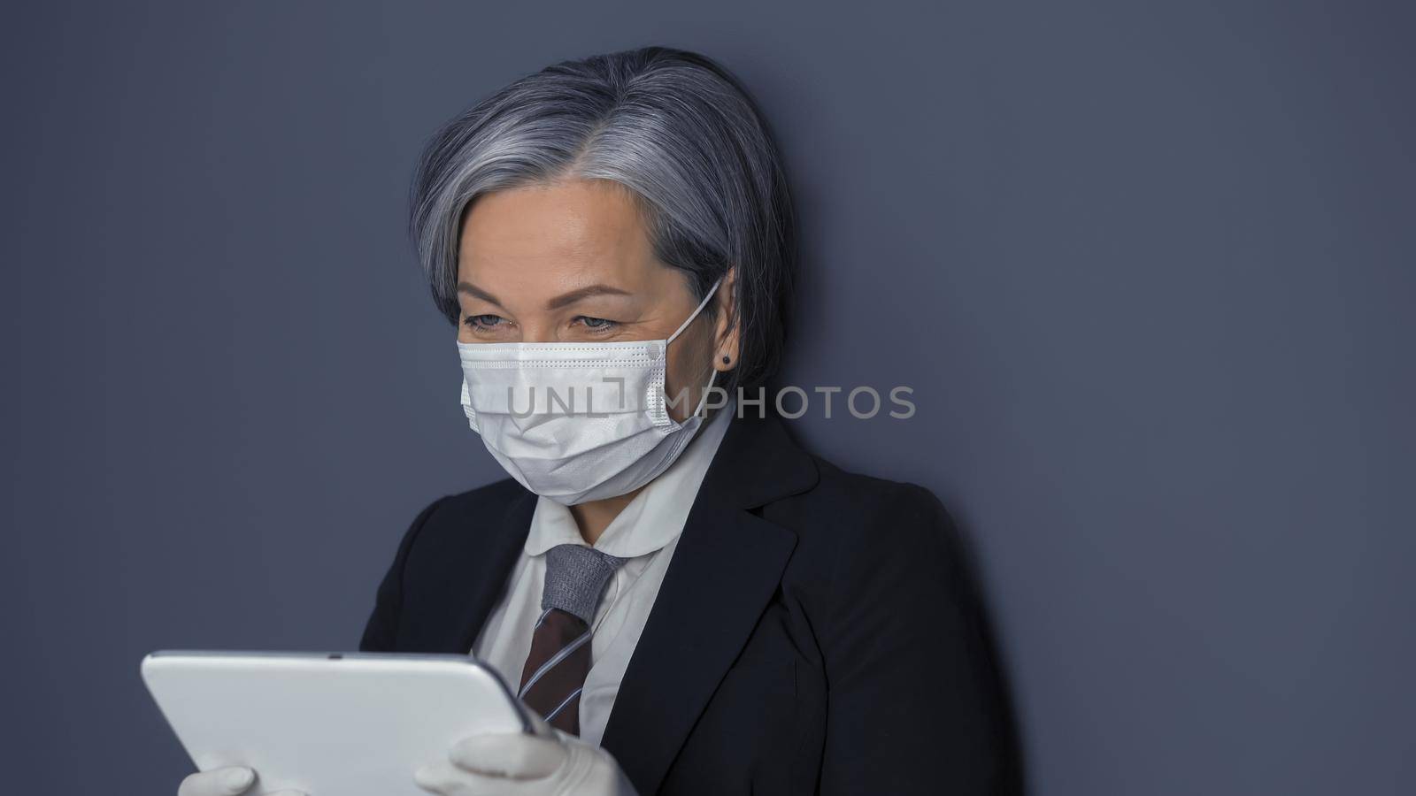 Mature businesswoman in protective mask using digital tablet. Gray haired woman reading news or working at tablet computer while standing on gray wall background. Toned image.