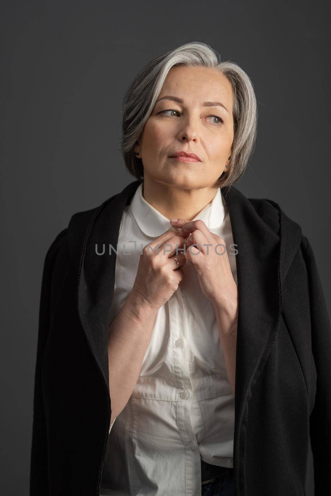 Charming gray-haired woman thoughtfully looks away adjusting collar of white shirt, throwing black jacket over her shoulders. Isolated on gray background. Lonely concept by LipikStockMedia