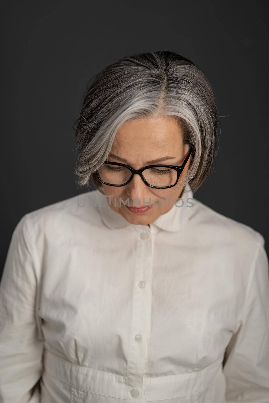 Lonely gray-haired mature woman looking down. Elegant depressed Caucasian lady in glasses and white shirt on gray background. Studio shot. Portrait.