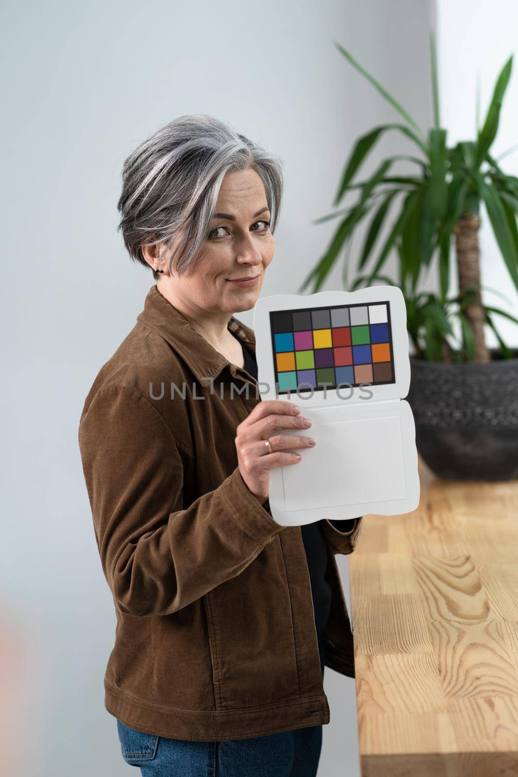 Pretty woman holds color checker or special target for color adjustment while standing in office interior. Gray haired woman showing color palette to adjust the color.