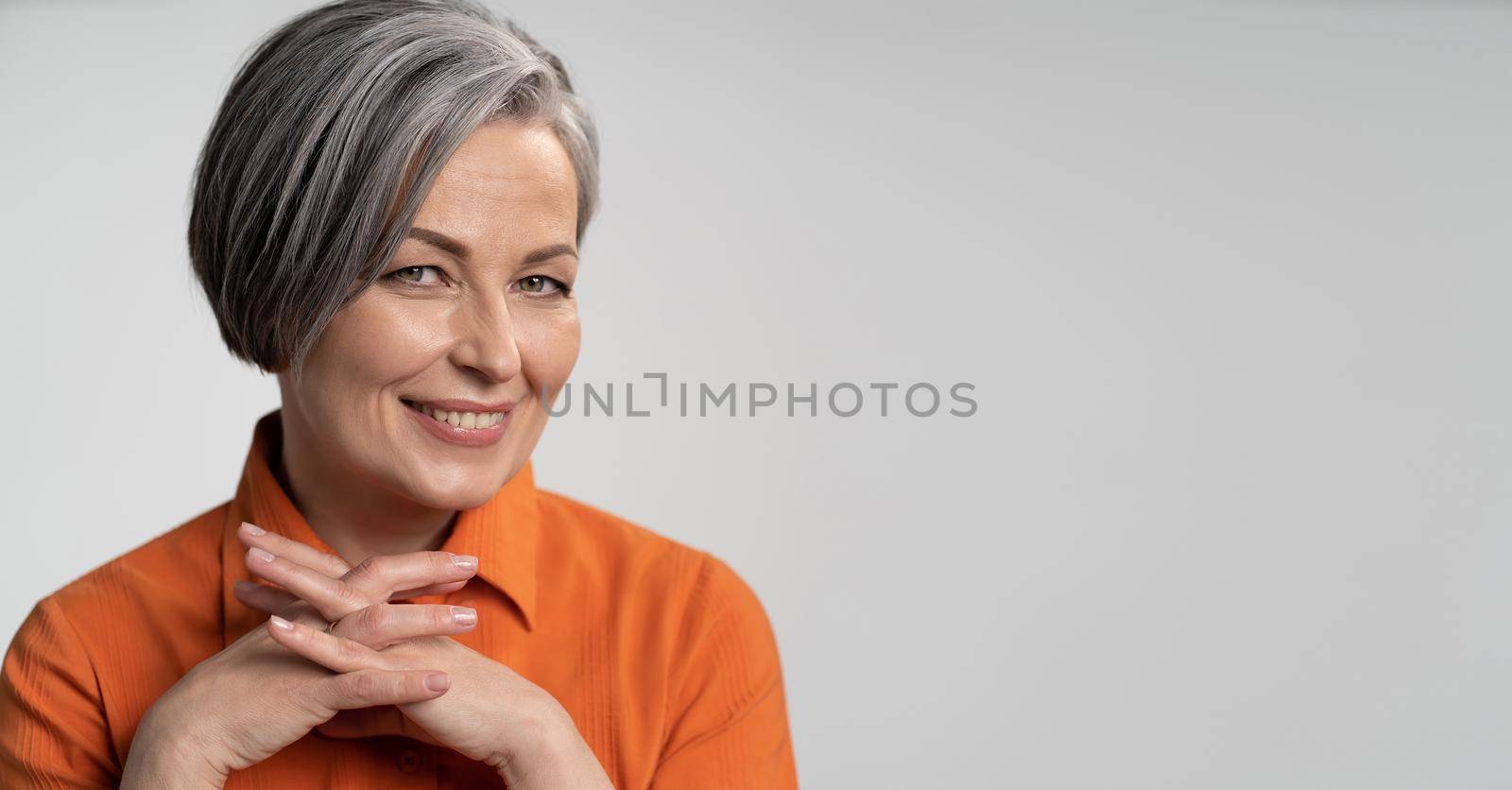 Happy aged woman smiling fingers crossed. Lady with bob hairstyle in orange blouse, head and shoulders portrait. Wisdom concept. Horizontal blank or template with copy spase at right side by LipikStockMedia