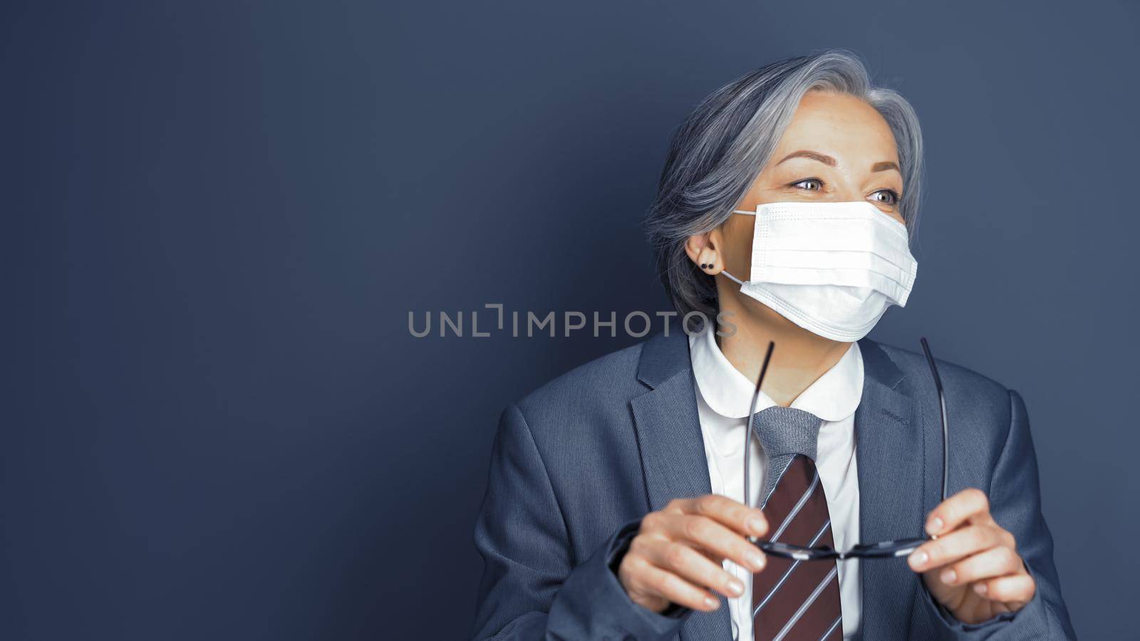 Businesswoman in protective mask holds eyeglasses looking aside. Pretty mature woman wearing business gray suit in motion on gray background with text space on left. Portrait. Toned image.