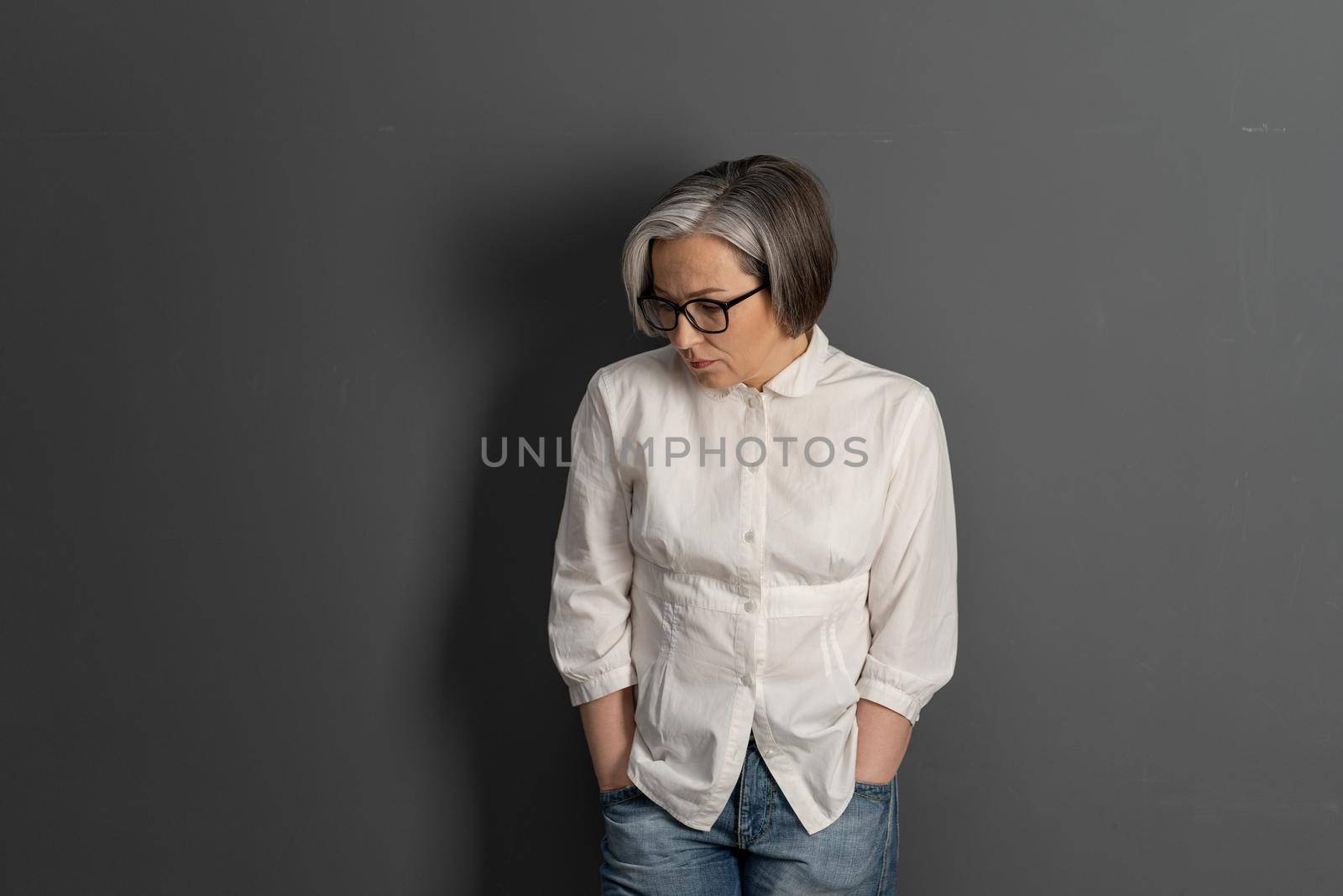 Sad middle-aged woman hunched looking down thoughtfully. Silver-haired stylish intelligent businesswoman with care hairstyle on grey background. Copyspace at both sides. Lonely concept.