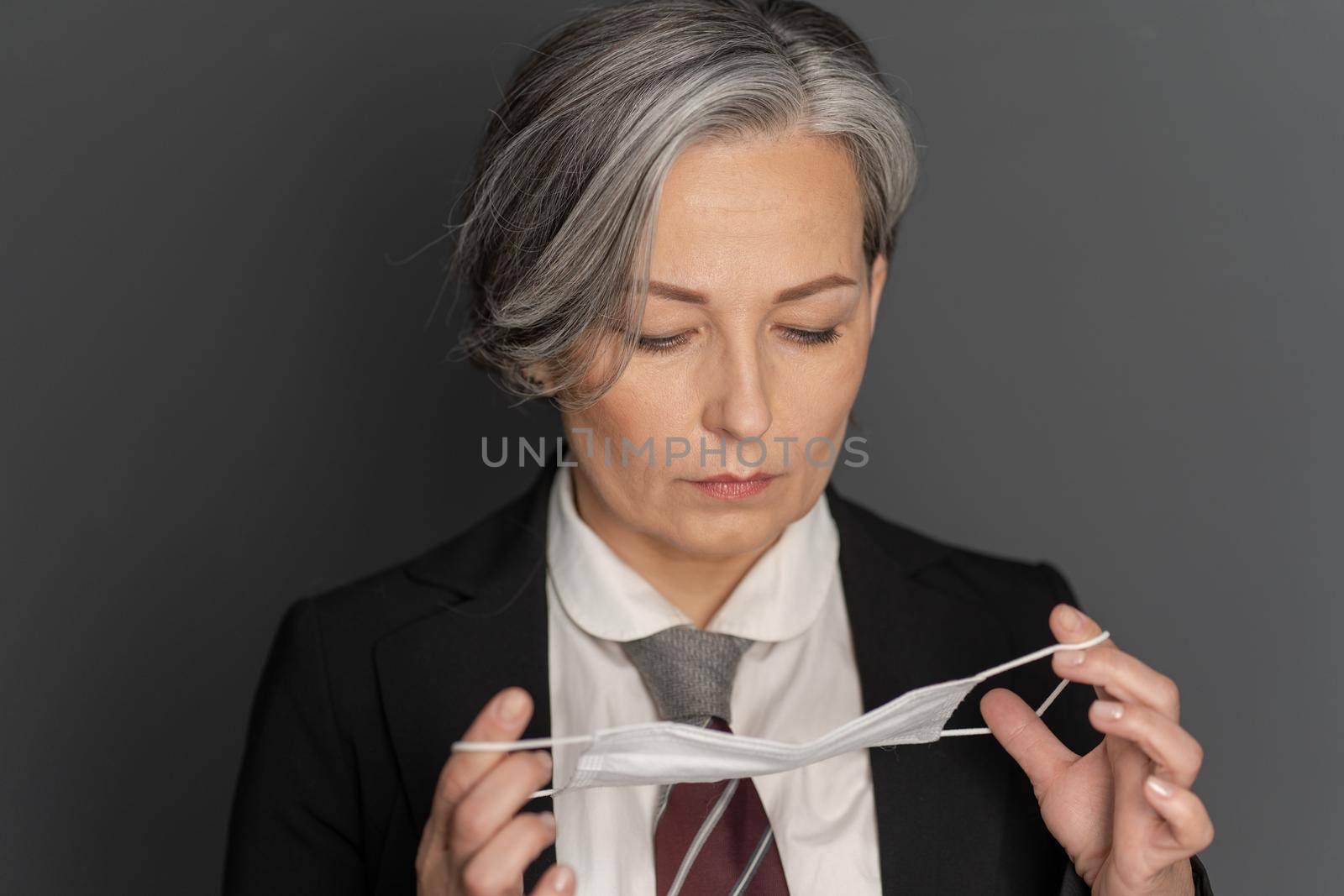 Pensive gray-haired business woman putting on a protective mask on her face looking down. Close up portrait. Pandemic concept. Protection concept.