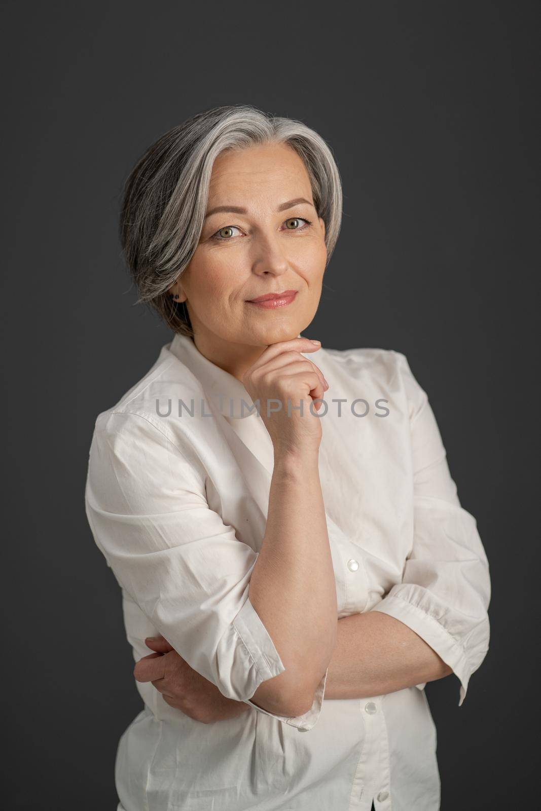 Beautiful gray-haired woman gently smiles while touching her chin with hand and looking at camera. Intelligent beautiful mature lady wearing white shirt standing alone on grey background.