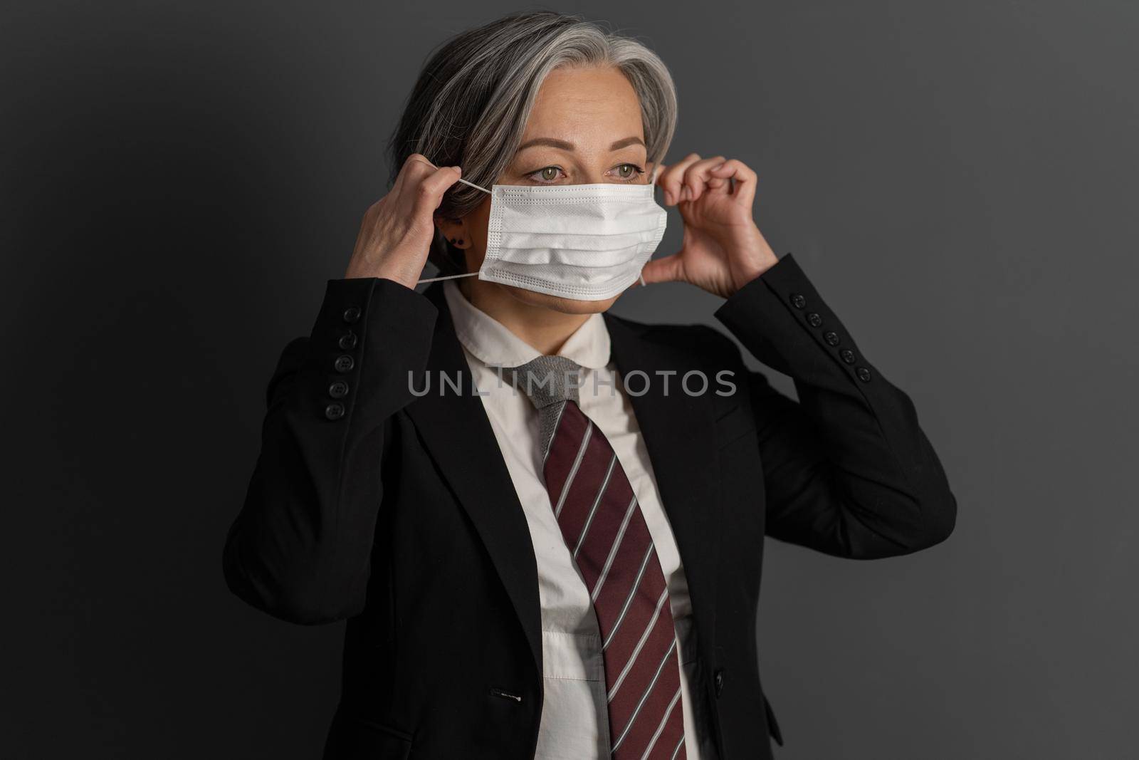 Pretty middle aged businesswoman putting on medical mask. Serious caucasian woman in black suit on dark gray background. Pandemic concept by LipikStockMedia