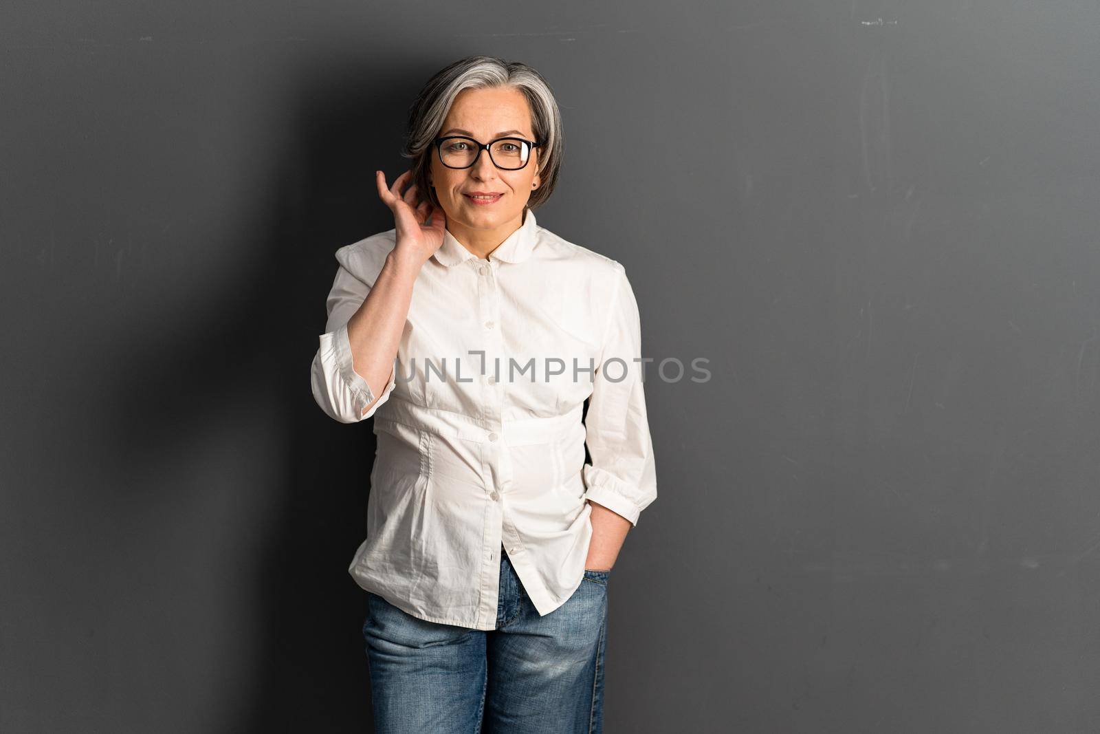 Pretty mature woman corrects medium-length hairstyle coquettishly. Adorable Gray-haired Caucasian model in jeans posing on gray background. Copy space or textspace at right side by LipikStockMedia