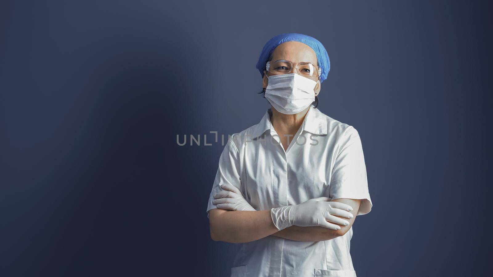 Female doctor or nurse in mask and white uniform standing crossed arms on blue gray background. Copy space for text at left side. Medicine concept. Tinted image.