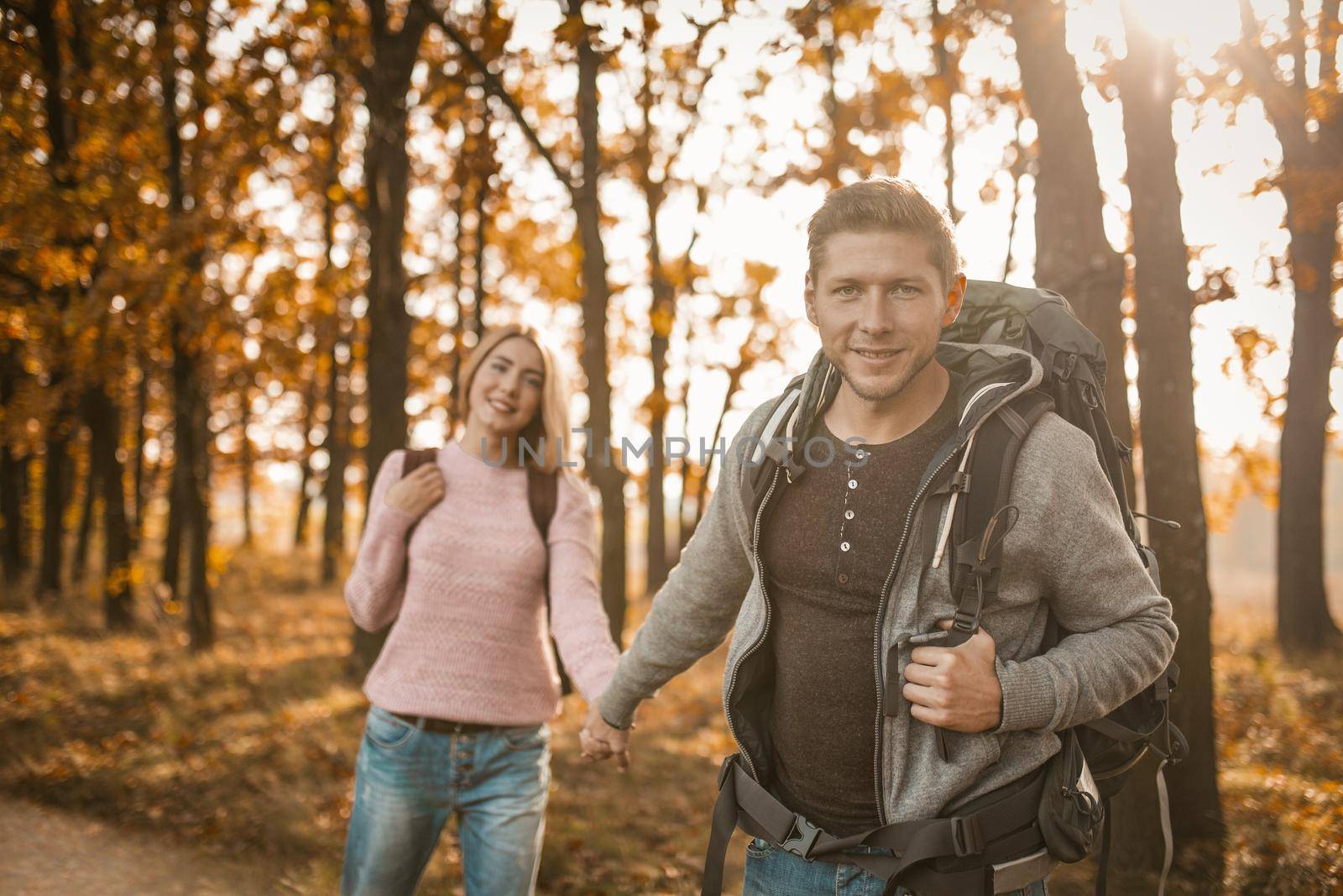 Two Travelers Walks Along Forest Trail Holding Hands, Selective Focus On Smiling Caucasian Man Looking At Camera While Standing In Back Sunlight, Guy And Girl With Backpacks Are Hiking Outdoors