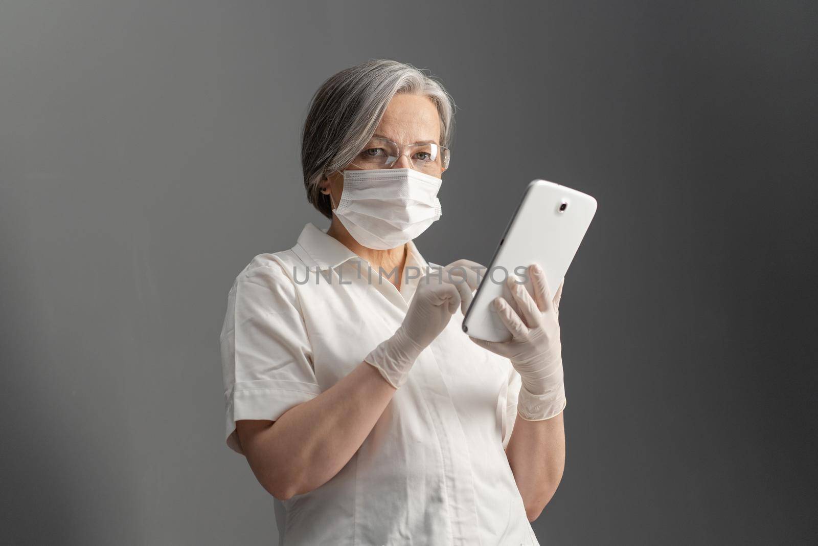 Serious female doctor works with tablet computer. Caucasian woman in white protective uniform uses digital tablet looking at camera while standing on gray wall background by LipikStockMedia