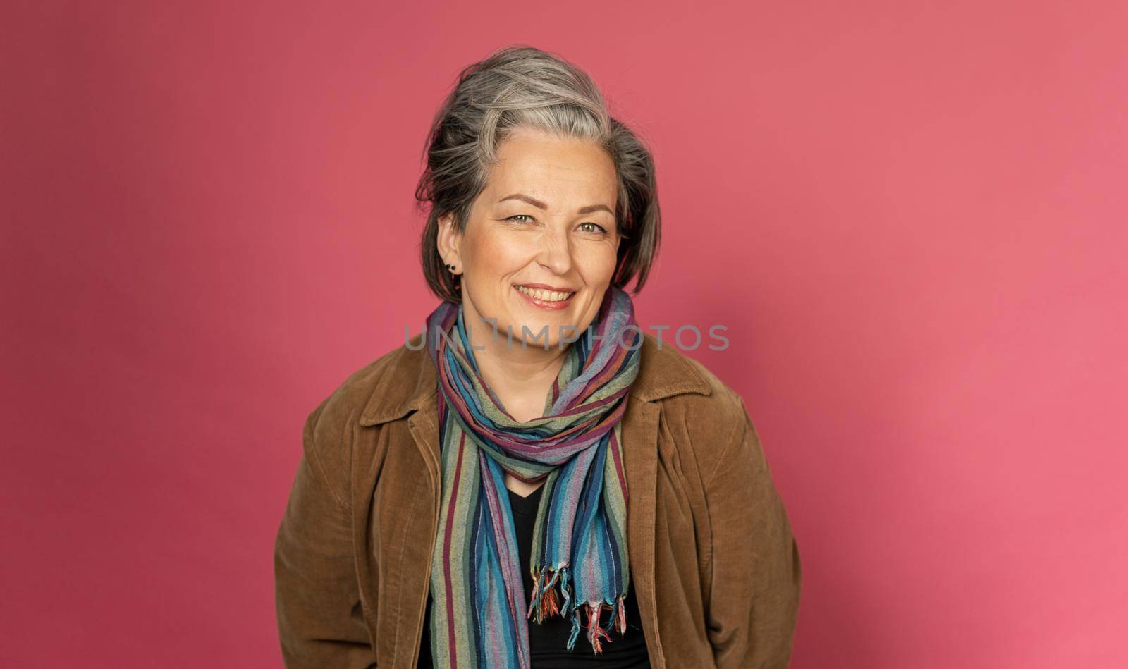 Happy gray-haired woman smiles broadly sexy posing in studio on pink background. Good-looking mature lady with nature make-up. Beauty concept. Vogue concept.