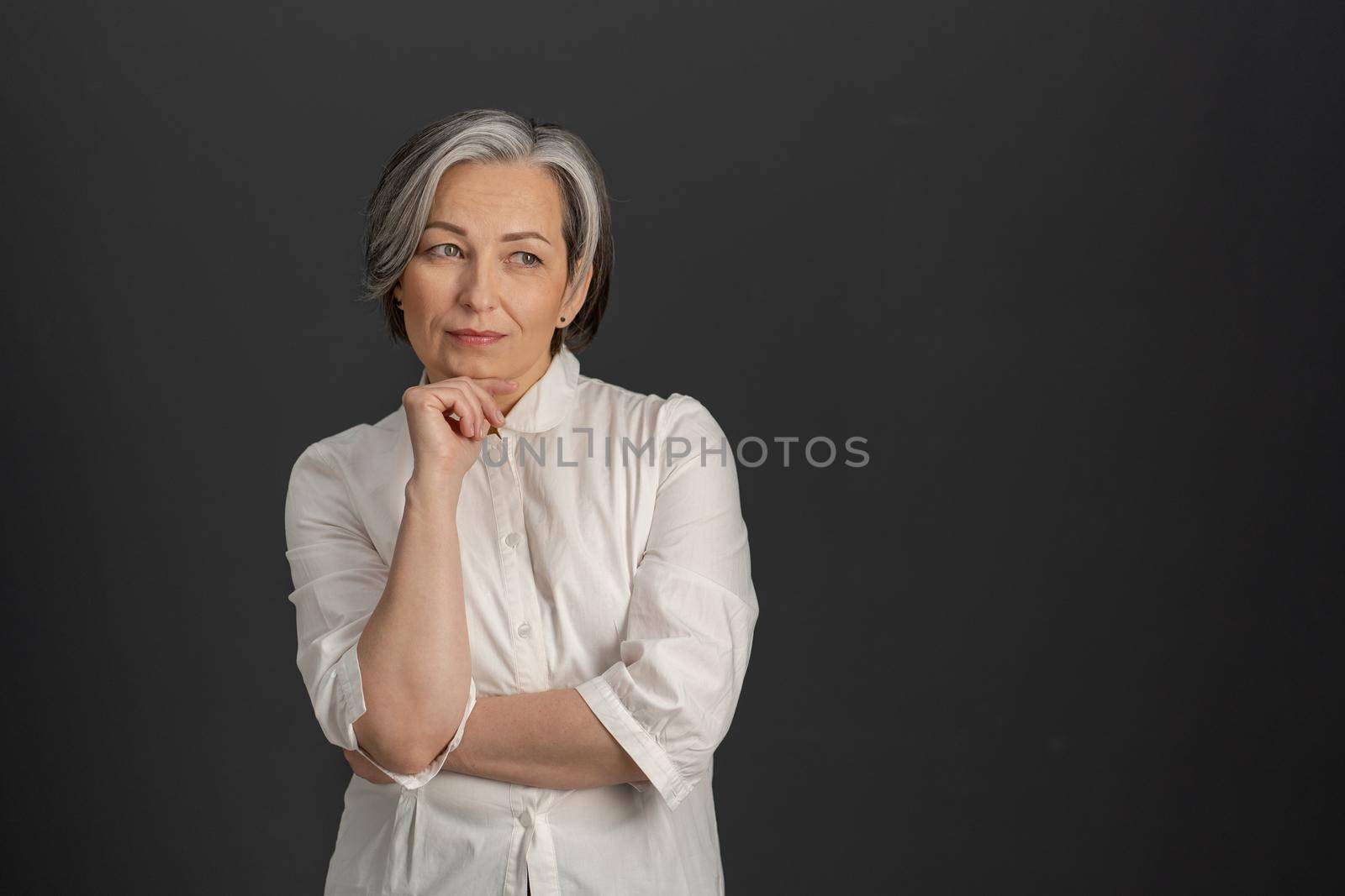 Thoughtful mature woman thinks touching by hand of her chin while looking at side. Gray-haired lady in white shirt isolated on gray background. Textspace at right side.