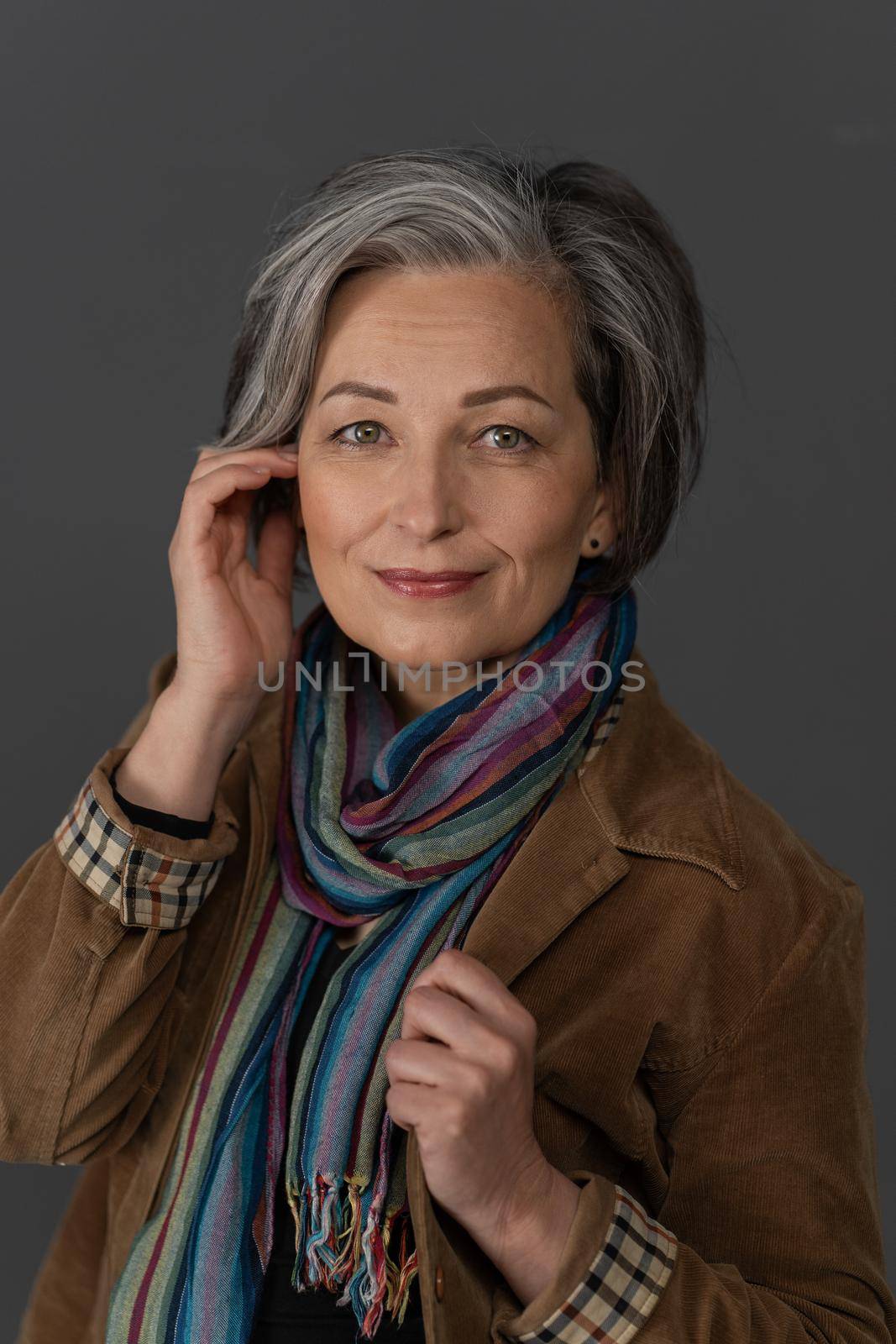 Stylish graying Woman charmingly smiles looking at camera. Caucasian mid aged beauty dressed in brown corduroy jacket with multi-colored scarf touches her face with hand. Anti-aging skin care concept.