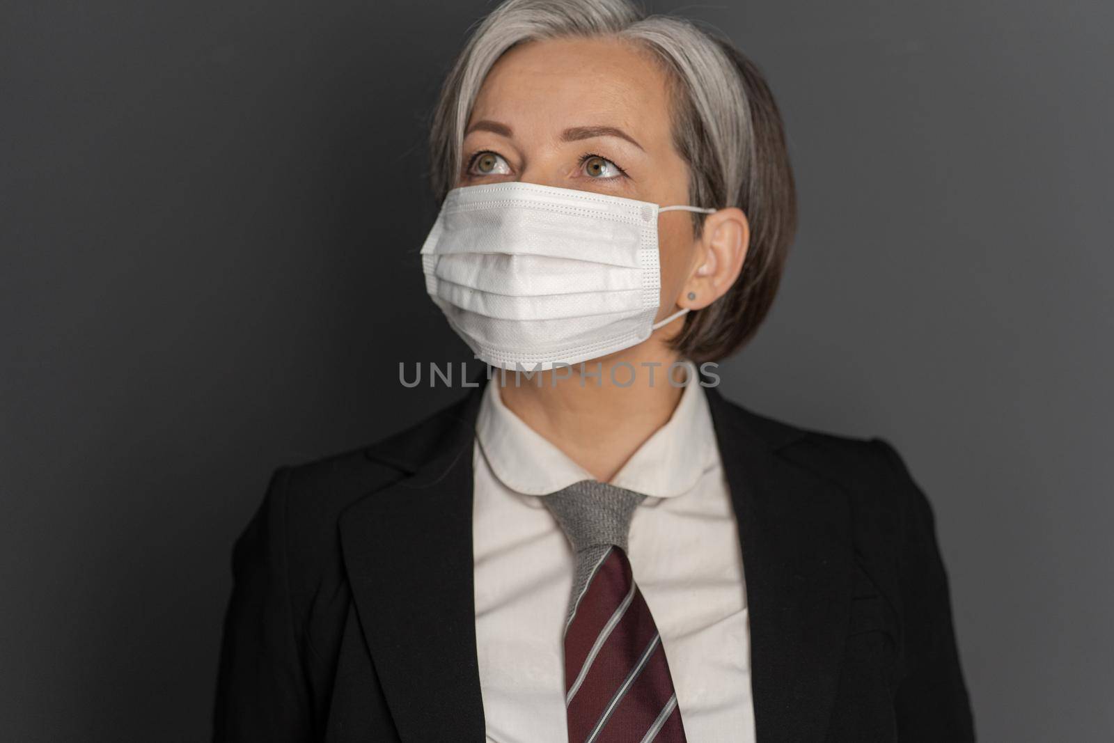 Gray-haired mid aged business woman in protective mask looking up. Selective focus on mask on female face. Virus outbreak concept by LipikStockMedia