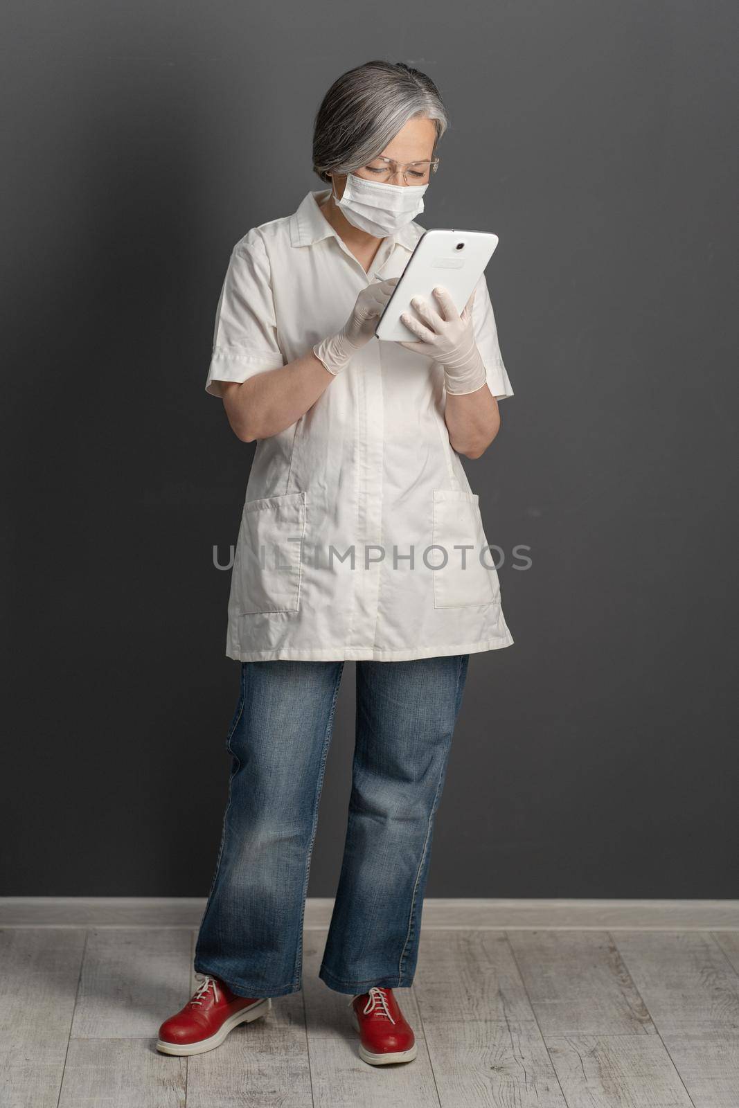 Female medic in mask and goggles using notepad. Mature gray-haired woman wearing in white coat, jeans and red boots standing full height. Medicare concept.