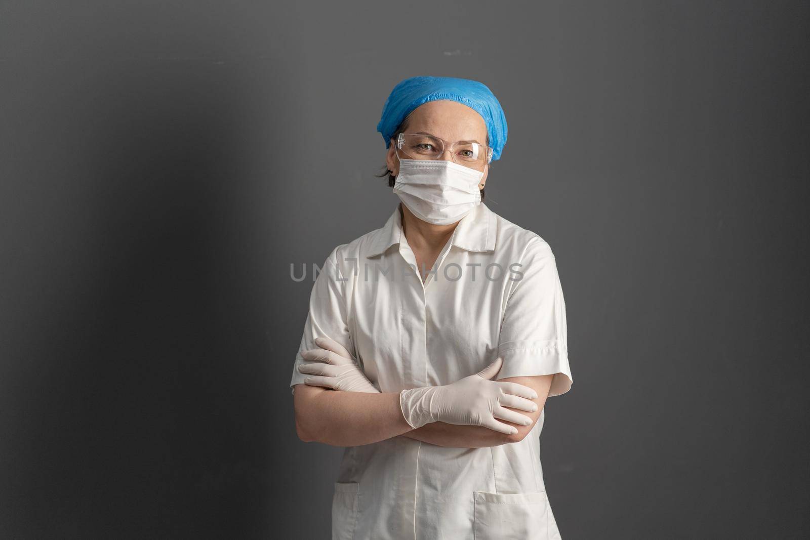 Positive doctor or nurse in protective uniform stands crossed arms on gray background. Caucasian female medic looks at camera. Textspace or copy space on left side.