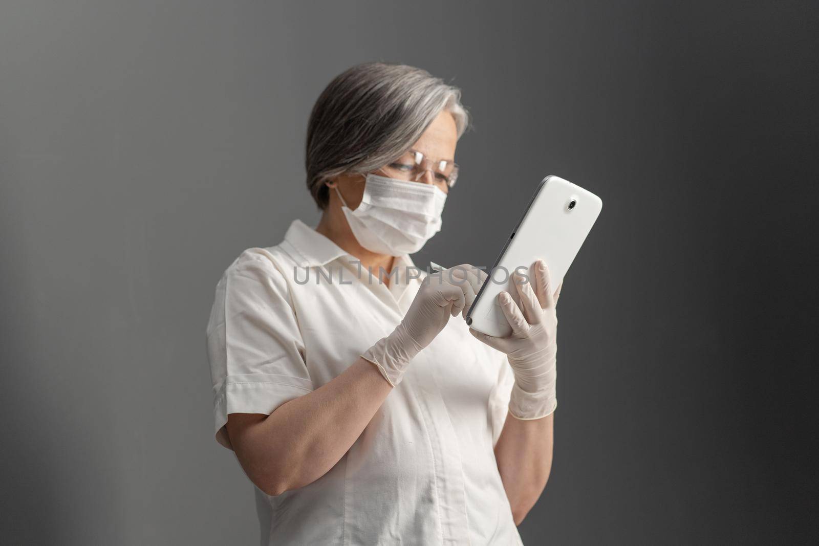 Side view of doctor using electronic tablet. Caucasian aged woman in protective mask works with digital gadget looking at screen on gray wall background. Focus on tablet in female hands.