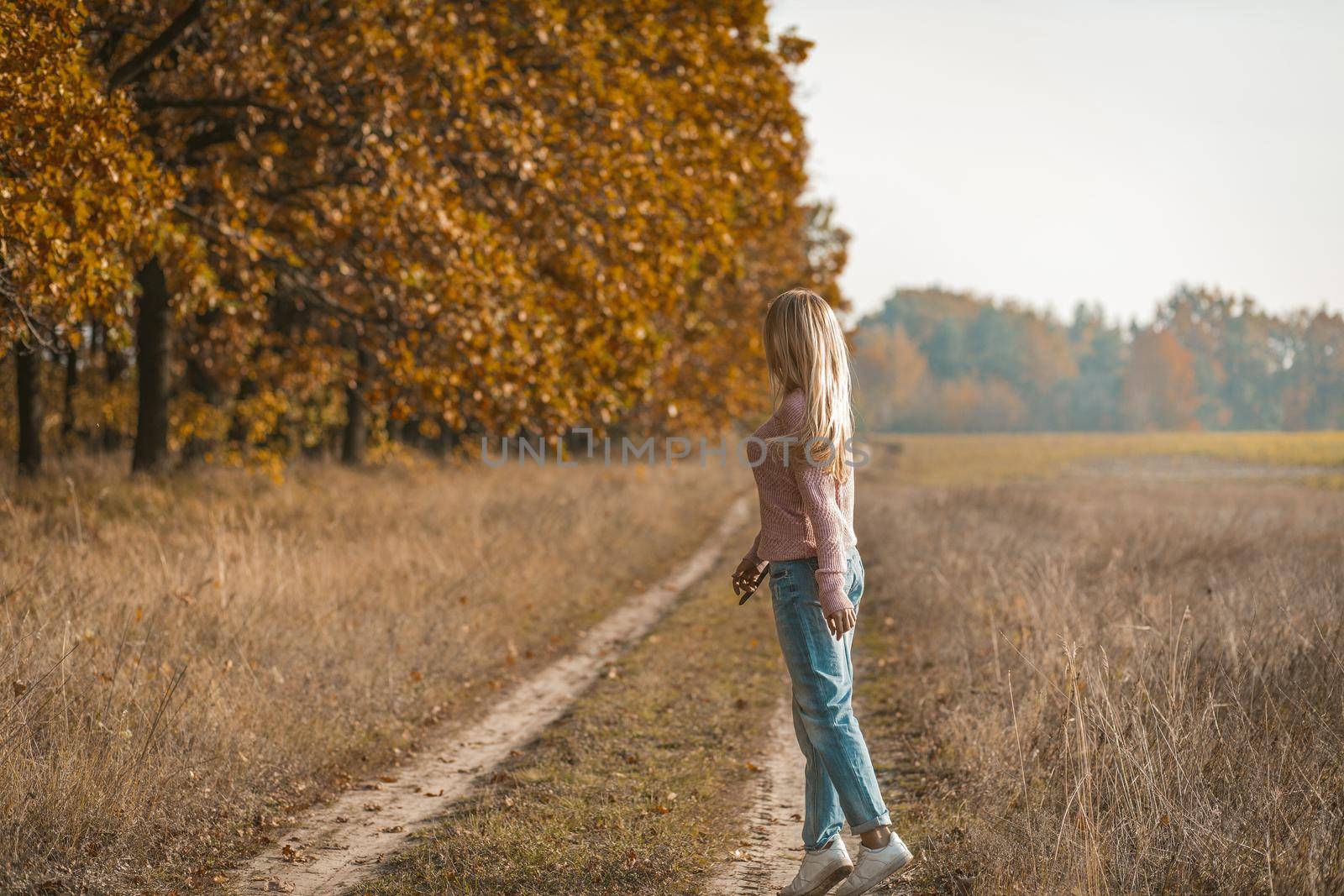 Rear view of a charming young blonde walking along a dirt road near the autumn forest, a young woman in casual clothes is tiptoeing in nature.