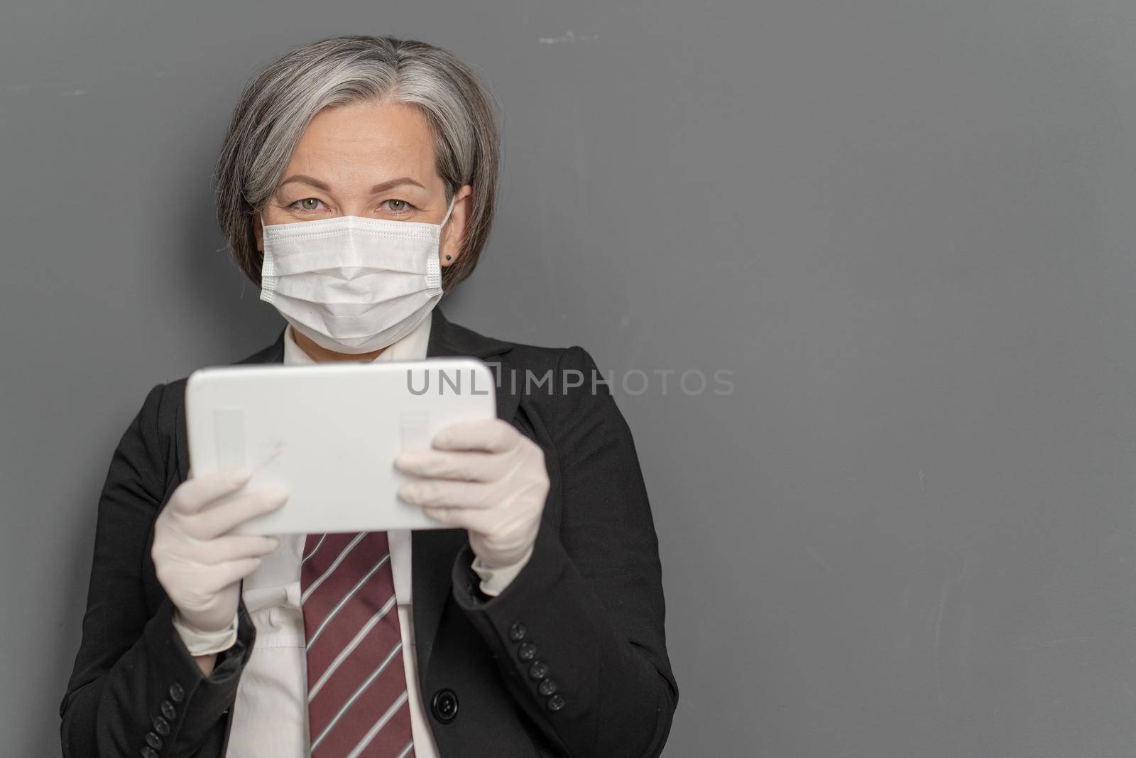 Gray-haired businesswoman works notepad. Caucasian lady holds tablet looking at camera on gray background with textspace on right. Focus on female face in protective mask. Virus outbreak concept.
