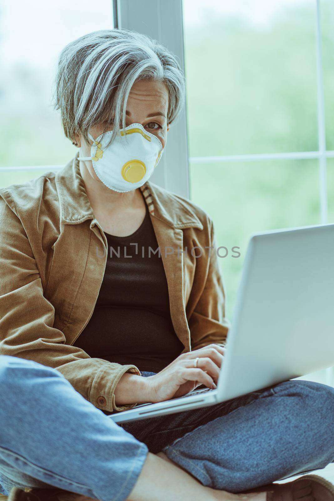 Gray-haired woman in mask fp1 looks at camera using computer while sitting tailor-fashion with crossed legs on a windowsill. Quarantine concept. Tinted image.