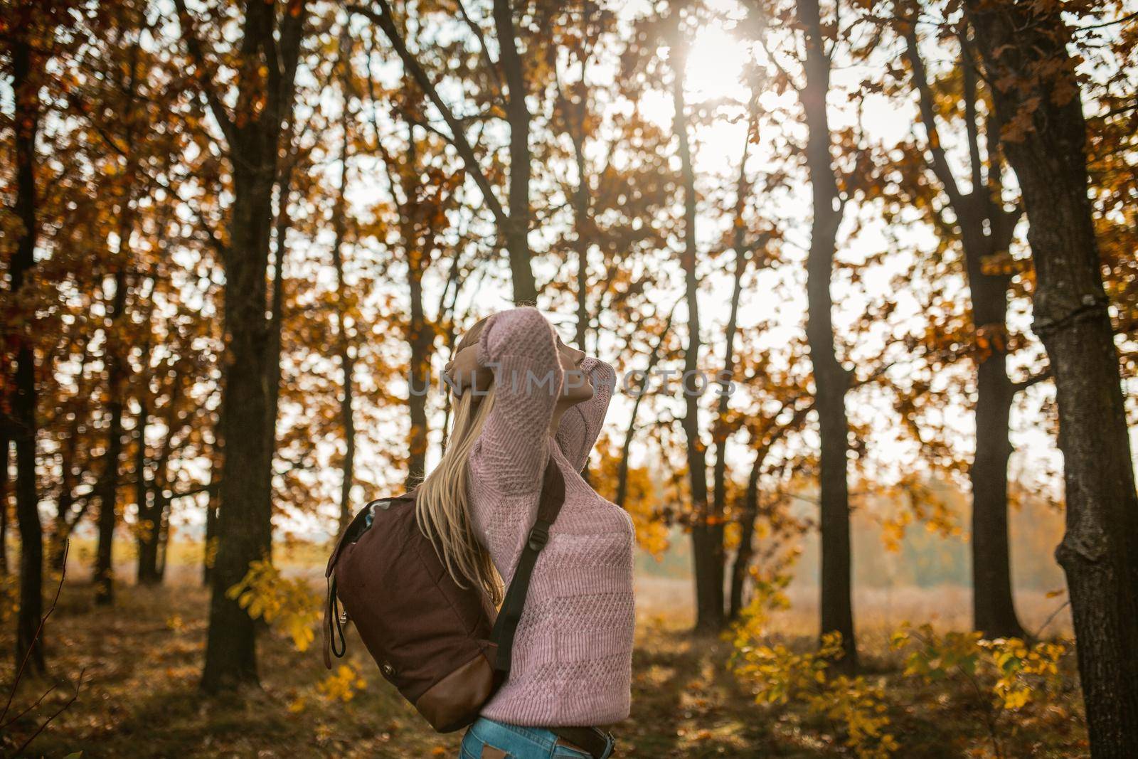 Tourist Woman Traveling In Nature, Young Caucasian Woman With Backpack Arched Her Back With Hands Behind Head And Set Face To The Sun While Standing Against Backdrop Of Autumn Trees On Sunny Day