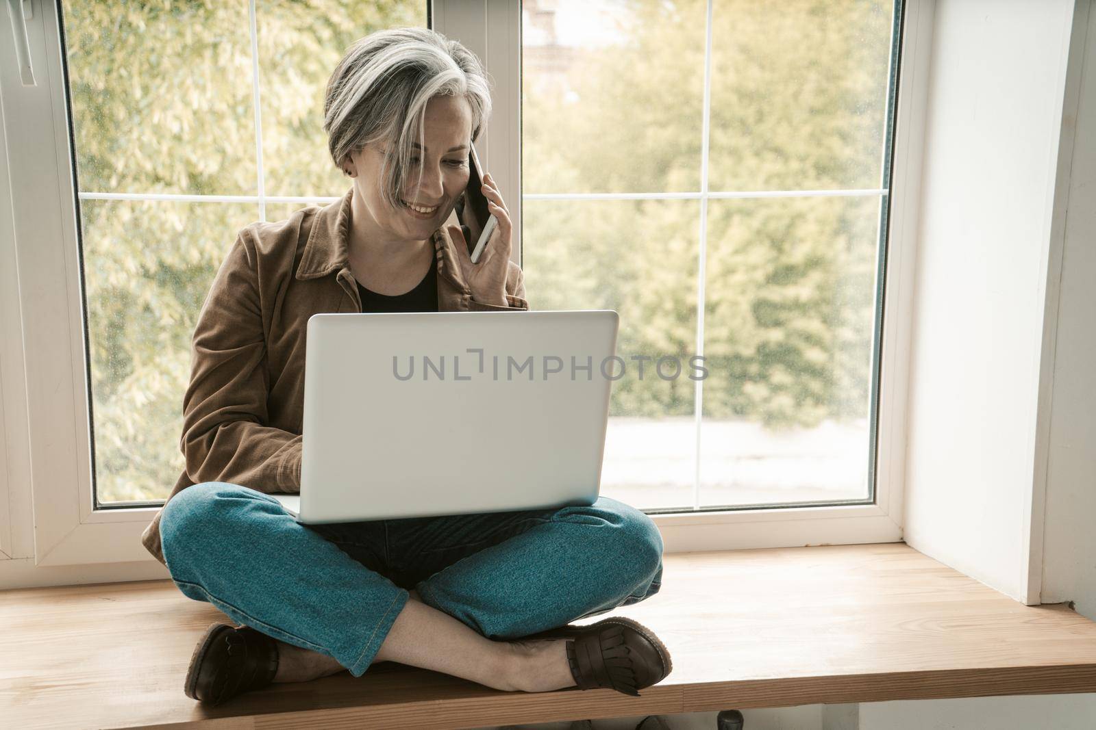 Mature freelance woman talks mobile phone working laptop computer while sitting tailor-fashion on wooden silll. Tinted image.