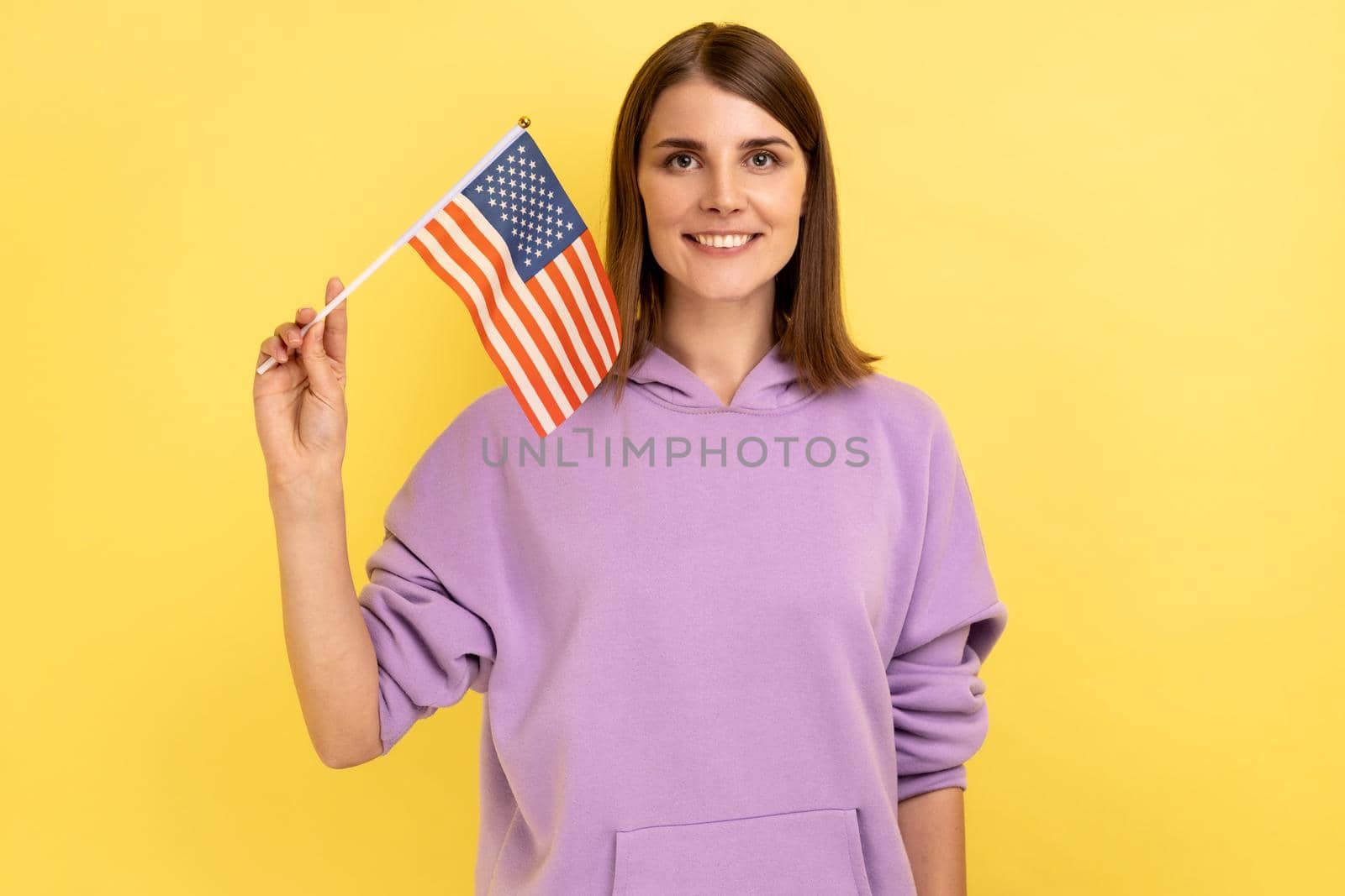 Portrait of delighted positive young woman standing and waving american flag, celebrating national holiday, wearing purple hoodie. Indoor studio shot isolated on yellow background.